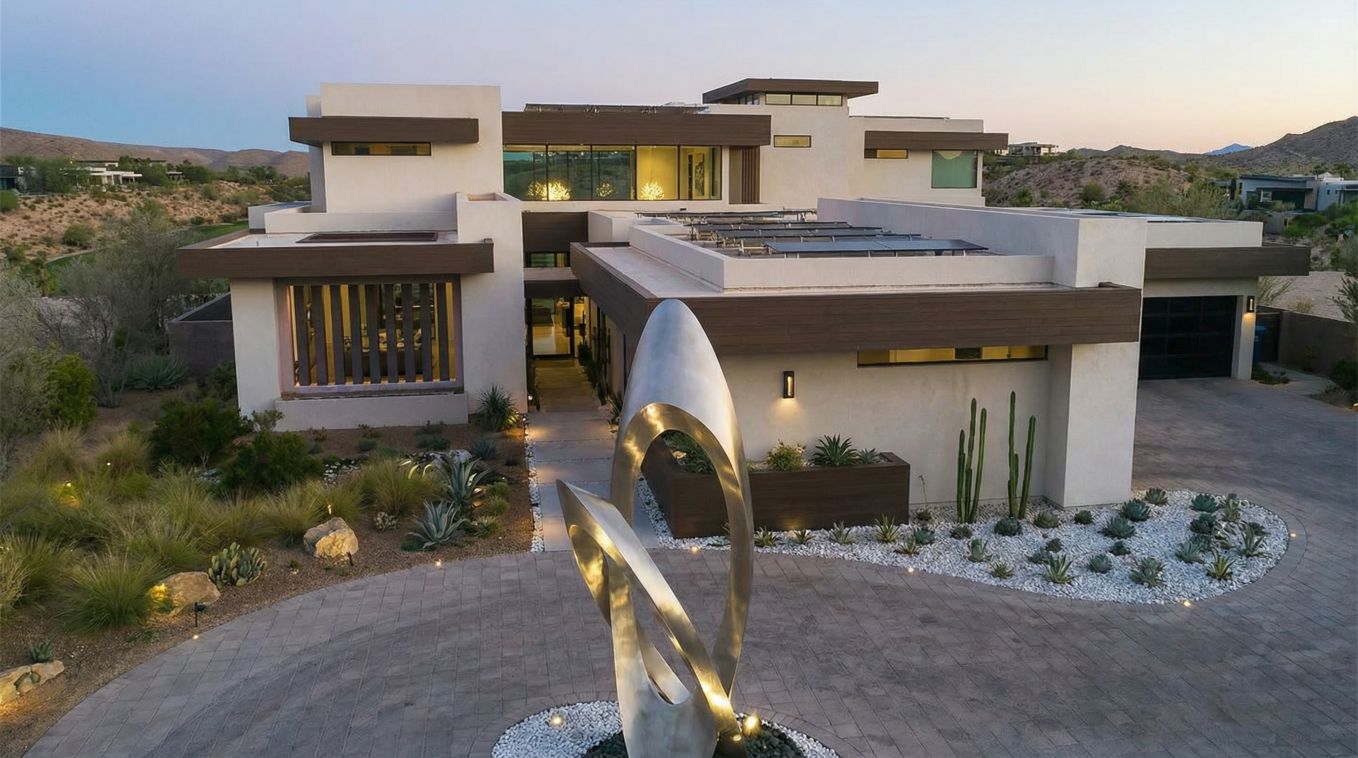 This image showcases the front exterior of a modern, multi-level home. The property features a desert landscaped yard with a circular driveway, a contemporary sculpture, and a neutral color palette with stone accents. The architecture emphasizes clean lines and geometric shapes, creating a striking and sophisticated entrance.