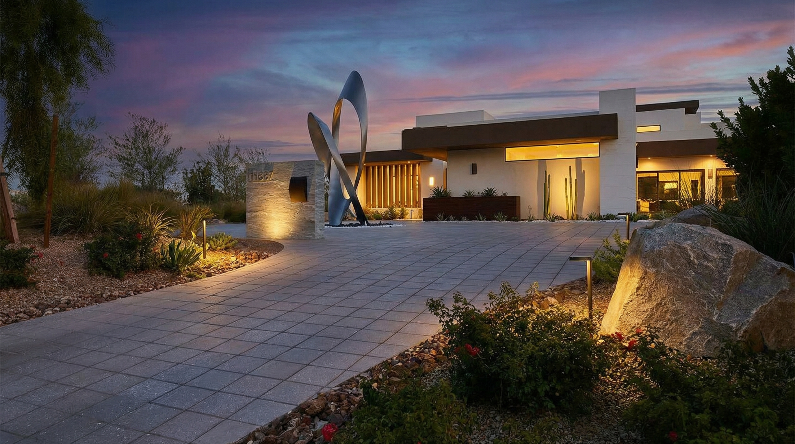 This is a striking front exterior view of a modern home at dusk. The property features a statement art piece in the front yard, complemented by professional landscaping, elegant walkway lighting, and a paved driveway. The architecture is contemporary, with clean lines and warm interior lighting visible through the large windows.