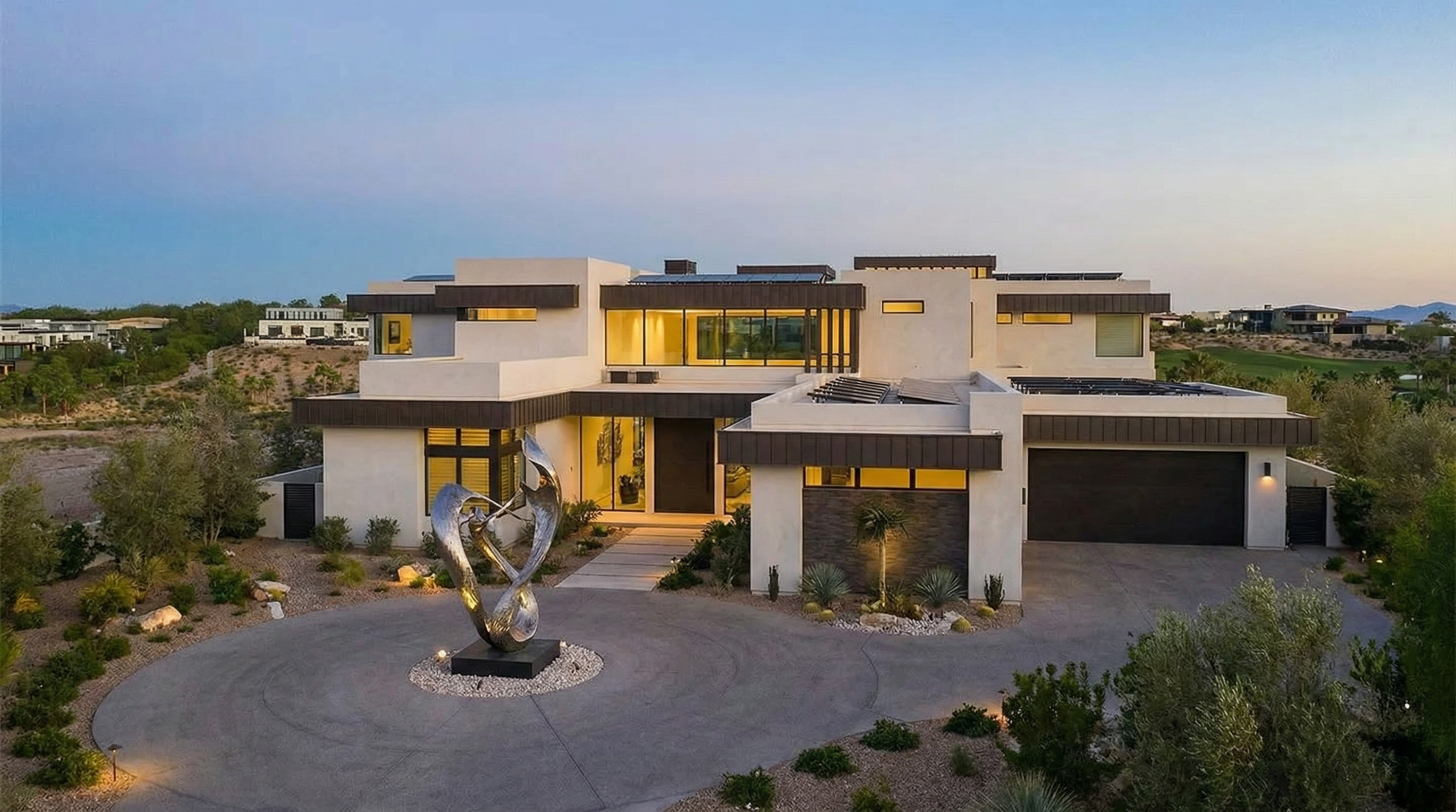This is a front exterior view of a modern luxury home. The residence features a multi-level design with a light-colored facade, dark-trimmed windows, and what appears to be solar panels on certain roof sections. A circular driveway leads to the entrance, and a contemporary sculpture is prominently displayed in the front yard, enhancing the property's curb appeal.