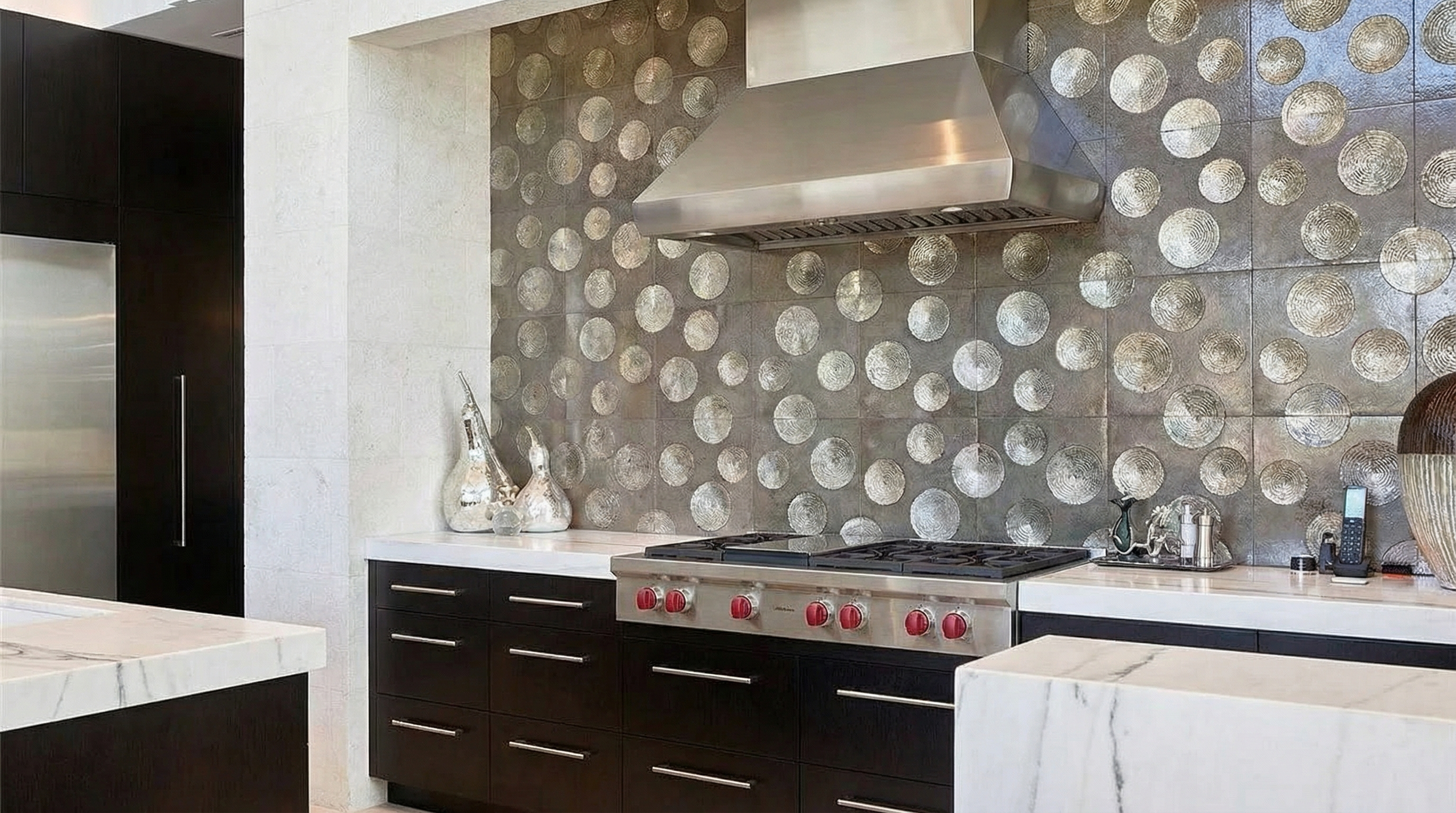This image showcases a modern kitchen area featuring dark wood cabinetry and a statement backsplash of circular patterned tiles. A professional-grade stainless steel range hood complements the gas cooktop below. White countertops provide a contrasting element, enhancing the sophisticated and functional design of the kitchen.