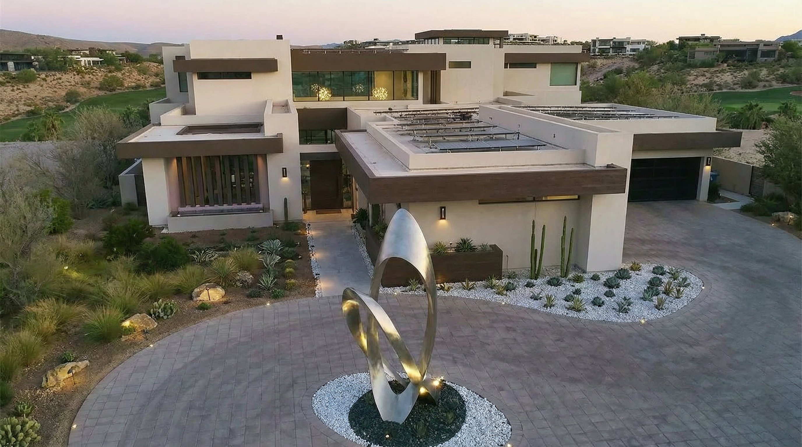 This image showcases the front exterior of a modern, luxury home. Featuring a striking sculpture in the driveway, the property boasts a clean, contemporary design with flat roofs and a neutral color palette accented by dark trim. Solar panels are installed on a portion of the roof, adding to the home's energy efficiency.