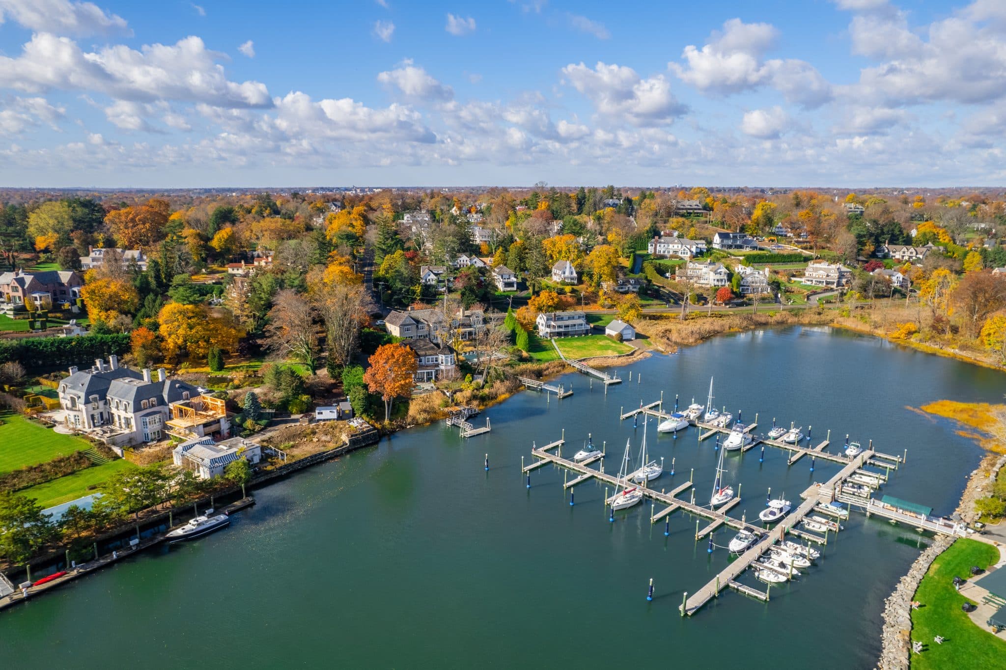 This scenic aerial view captures a luxurious waterfront property situated along a tranquil riverbank, featuring a large estate home and a private deep-water dock with several docked boats. The surrounding landscape is characterized by vibrant autumn foliage, lush green lawns, and other beautiful residential homes dotting the shoreline. The image conveys an affluent, serene lifestyle with a strong emphasis on water access and seasonal natural beauty.