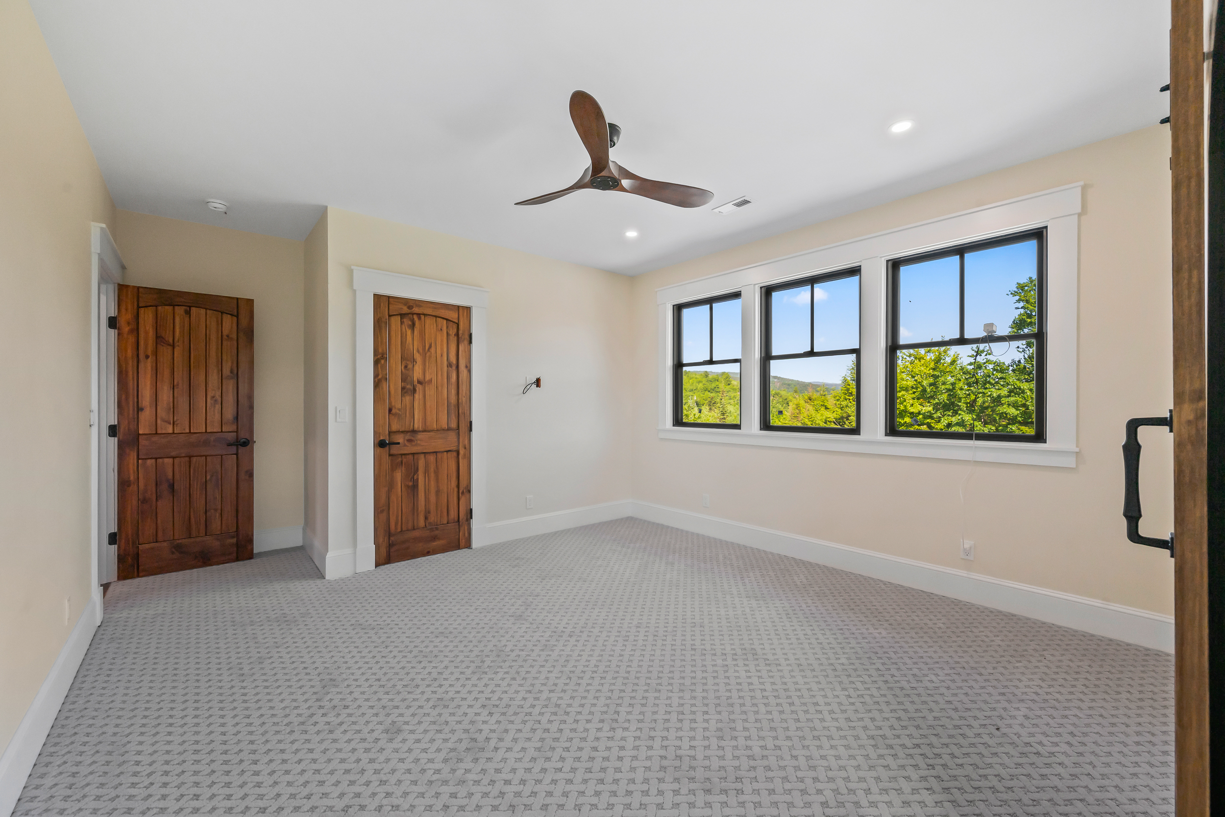 This spacious, unfurnished bedroom features neutral-toned walls and unique patterned carpeting that adds texture to the room. Three large windows offer abundant natural light and scenic views of the surrounding trees and hills, while a modern wooden ceiling fan serves as a central focal point. Two classic wooden doors with traditional trim highlight the residence's careful attention to finish and quality craftsmanship.