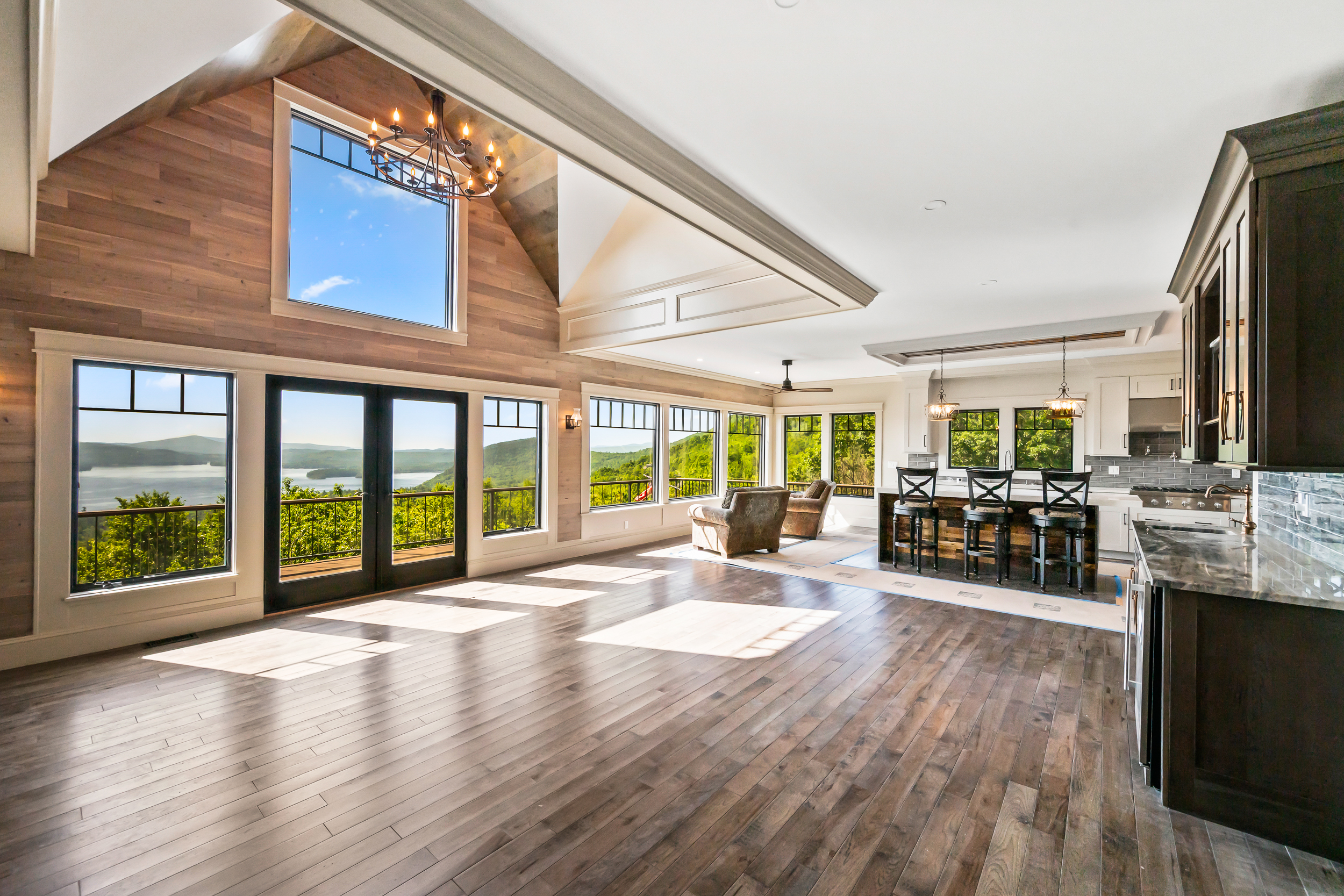 This spacious, open-concept living area seamlessly integrates a gourmet kitchen with a grand living space, highlighting stunning mountain and lake views through expansive windows and double doors. Rich hardwood flooring flows throughout, complemented by a tall, vaulted wood-paneled ceiling and a rustic-modern chandelier. The aesthetic leans toward a sophisticated mountain lodge style, emphasizing natural light and scenic beauty.