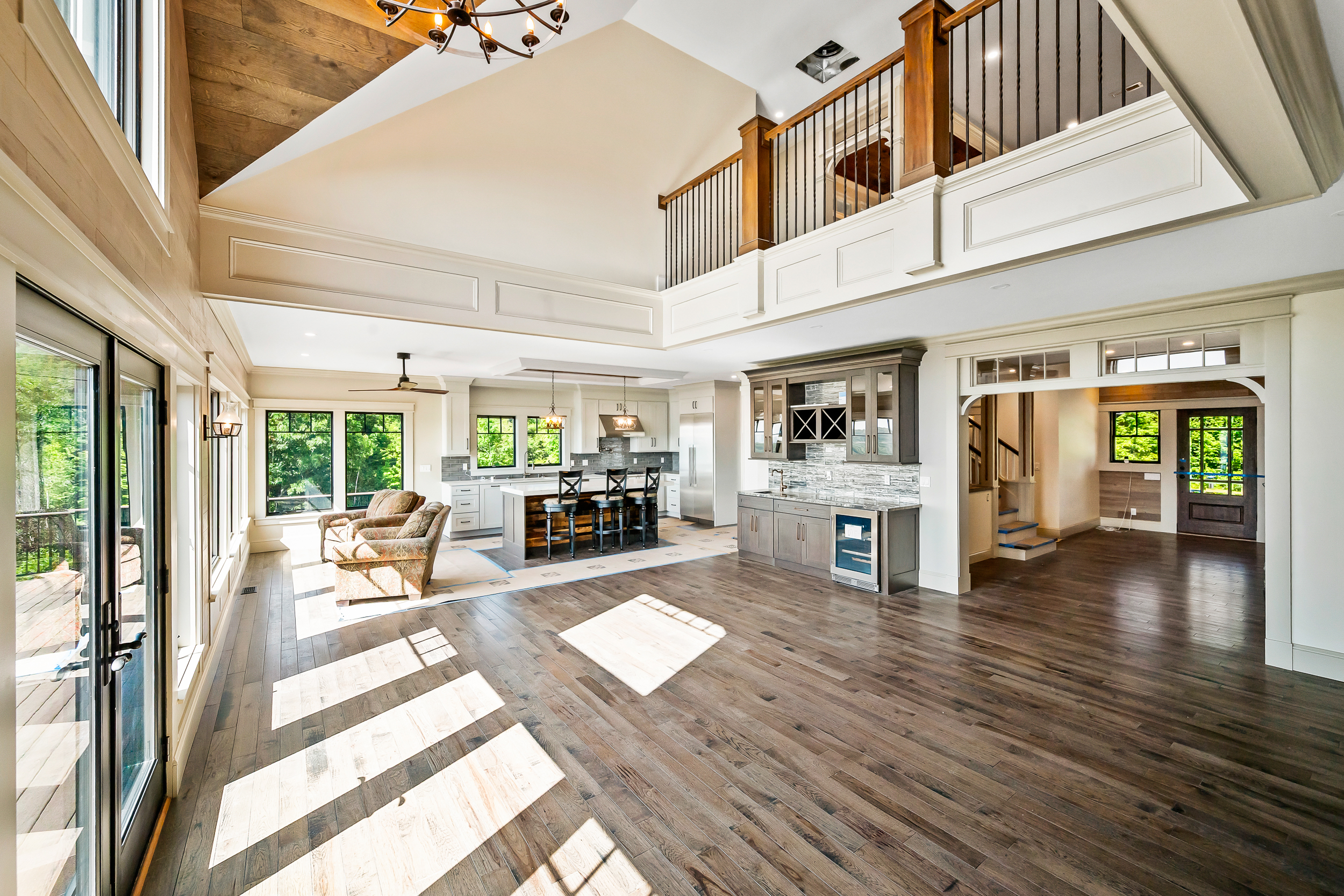 This spacious, open-concept living area features stunning vaulted ceilings and a dramatic lofted balcony, creating an airy and grand atmosphere. The room boasts beautiful dark hardwood floors, a sophisticated wet bar area with a wine cooler, and seamless transitions into a modern kitchen, making it perfect for entertaining. Large windows and glass doors flood the space with natural light while providing picturesque views of the lush outdoors.