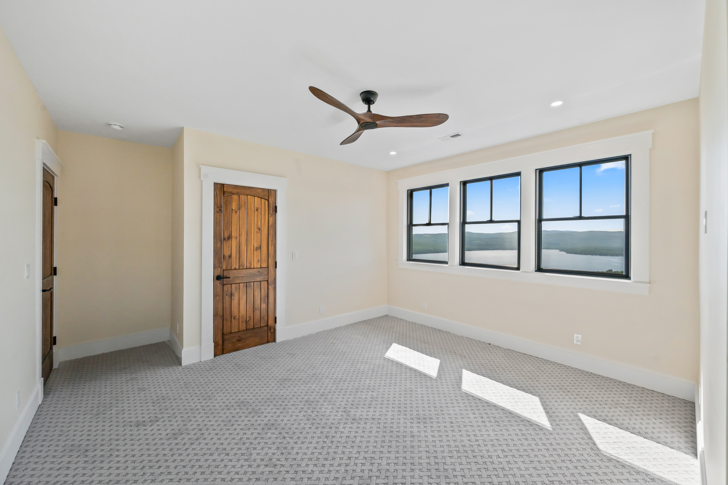 This spacious, unfurnished bedroom features neutral wall tones and a textured, patterned carpet that creates a clean, inviting atmosphere. A prominent wooden door with a rustic design adds character, while three large windows offer a stunning, expansive view of a lake and mountainous landscape. The room is further enhanced by a modern ceiling fan and recessed lighting, making it a bright and versatile space.