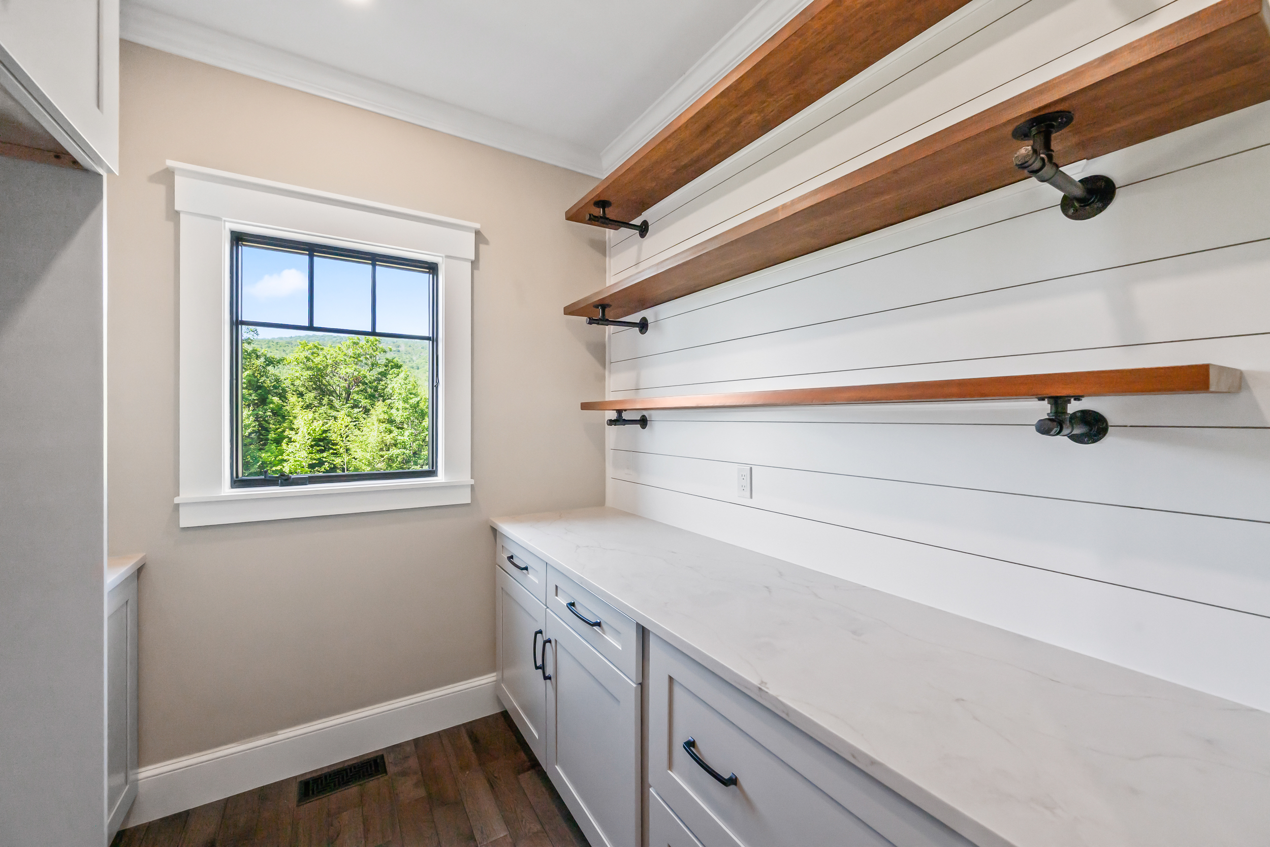 This clean and well-lit pantry or butler's pantry features beautiful shaker-style cabinets topped with a white marble-look countertop. Open shelving with industrial-style iron pipe brackets enhances the clean, farmhouse-inspired wall paneling. A large window provides natural light and a pleasant view of the woods, making the space feel both functional and inviting.