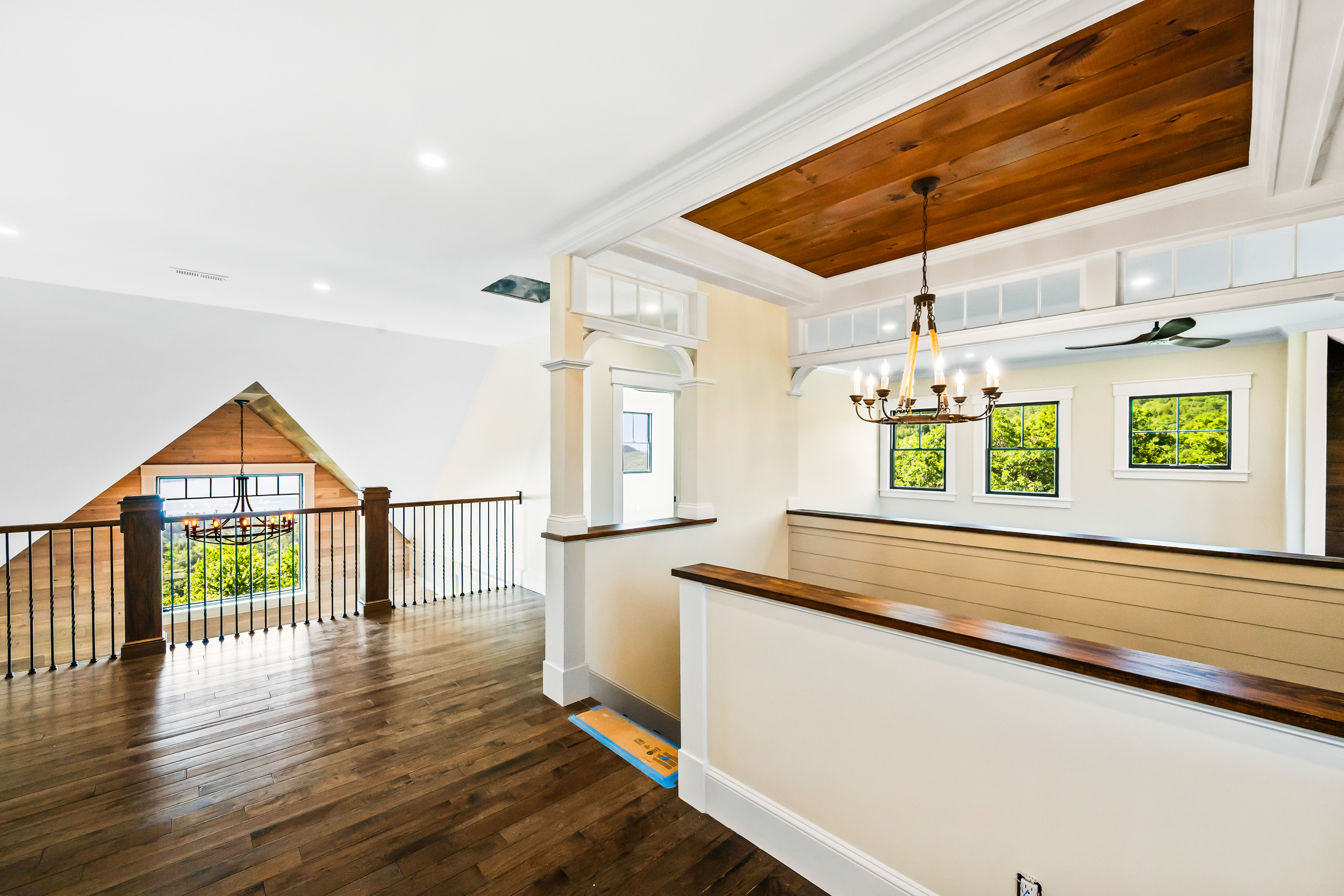 This spacious, light-filled hallway features rich hardwood flooring, a unique wood-paneled ceiling accent, and traditional railing details. The open design connects different levels of the home with elegant architectural touches like transom windows and views of the surrounding woods, creating a welcoming and upscale atmosphere.