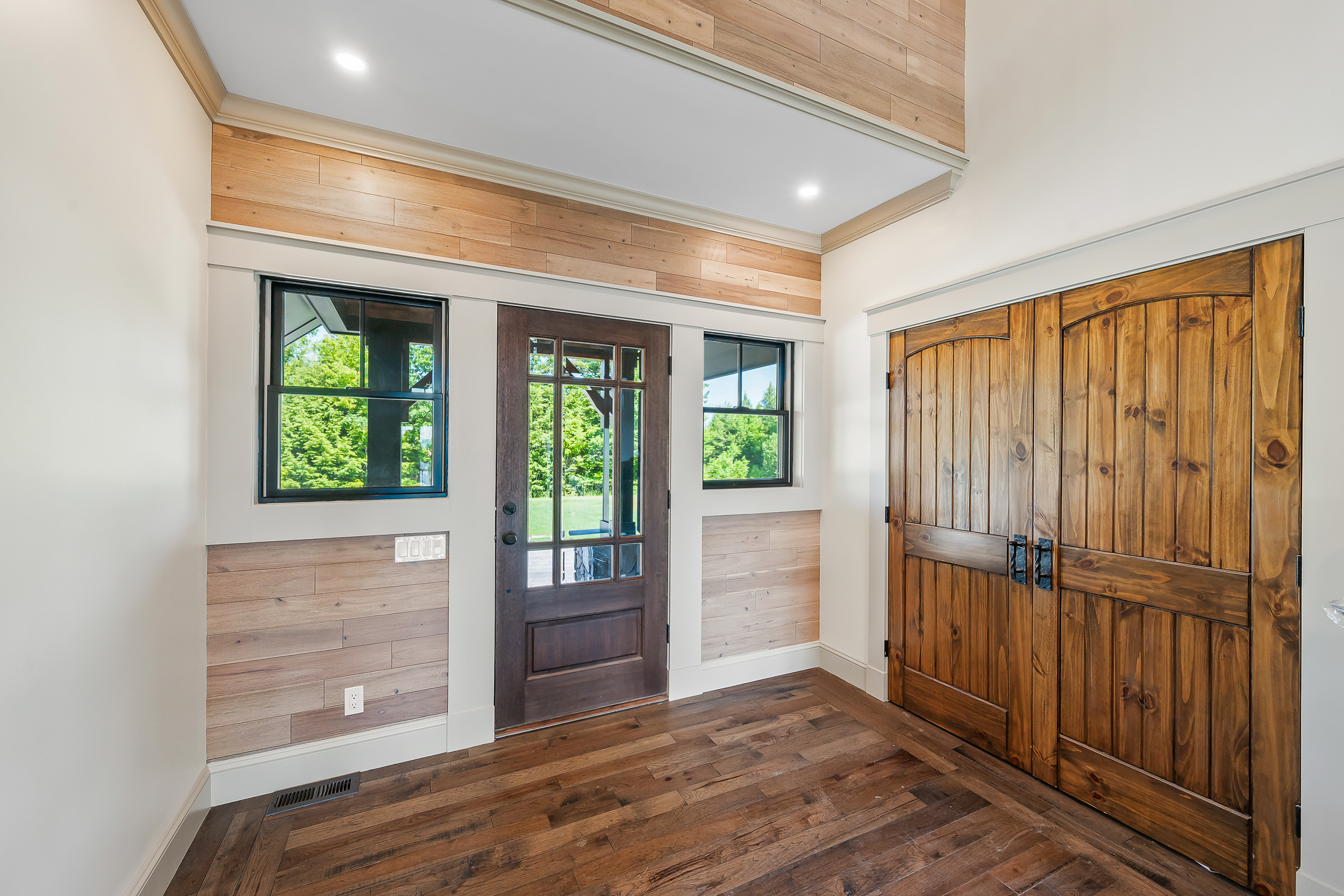 This bright entryway features a blend of farmhouse and modern aesthetics with rustic wood planking accent walls and a rich dark wood front door. The space includes elegant double barn-style doors leading to a closet and high-quality dark hardwood flooring throughout. Large, dark-framed windows frame the front door, allowing ample natural light to flood the inviting, refined living space.