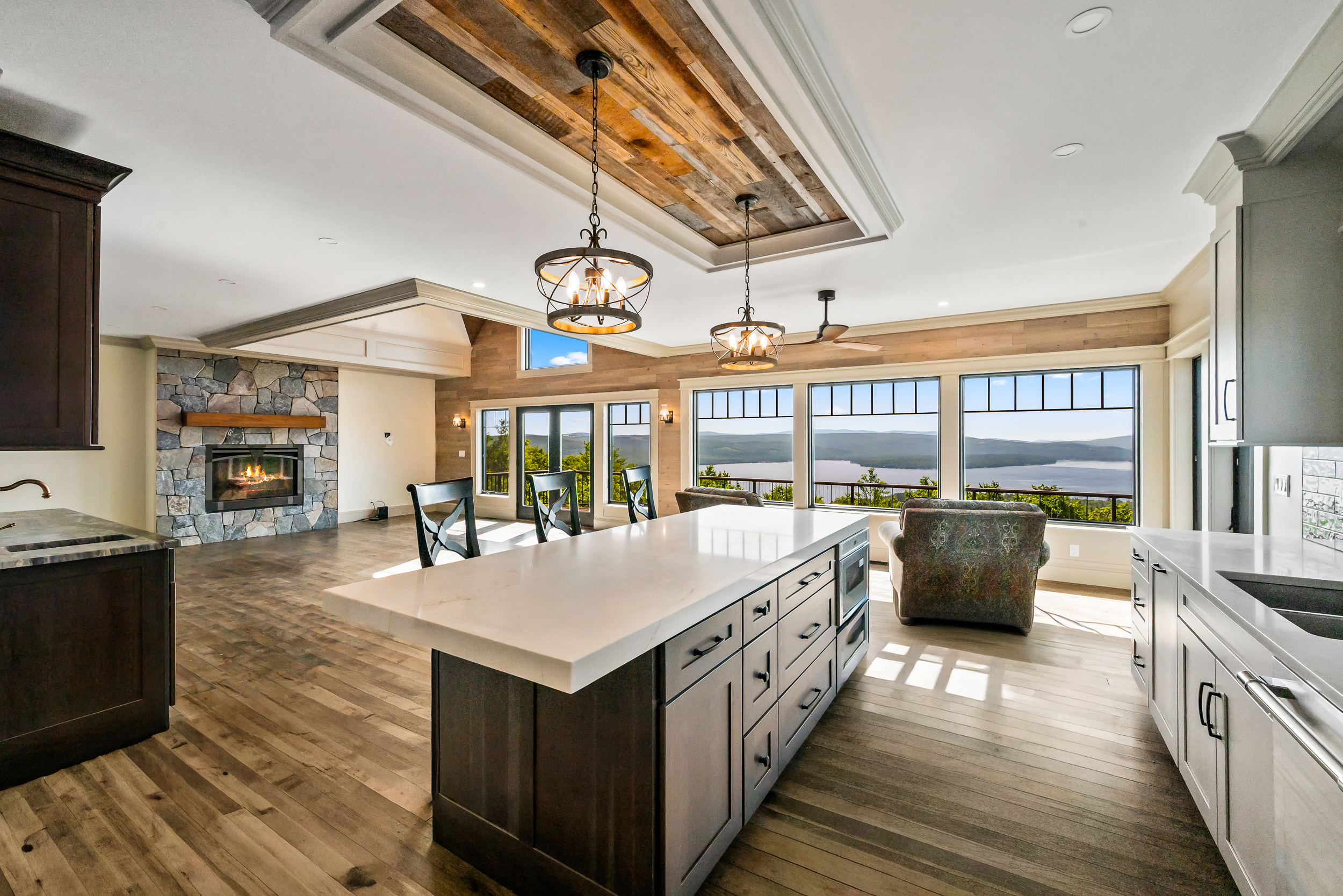 This stunning open-concept kitchen features a large center island with a sprawling light-toned quartz countertop, dark wood cabinetry, and elegant industrial-style pendant lighting. The space seamlessly integrates with a living area boasting a magnificent stone fireplace and floor-to-ceiling windows that provide panoramic views of a lake and mountainous landscape. With its blend of rustic reclaimed wood ceiling details and modern finishes, this home offers a luxurious yet cozy mountain-retreat aesthetic.