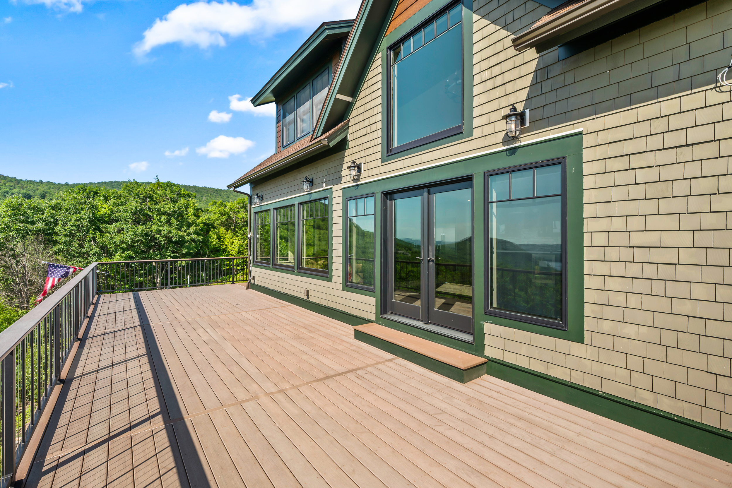 This image features a spacious, composite-material deck wrapping around the side of a modern mountain home. The exterior is finished in attractive olive-green shingles with dark-framed windows and doors, offering a clean, contemporary aesthetic. A sleek metal railing ensures safety while providing unobstructed views of the surrounding lush forest and mountain horizon.