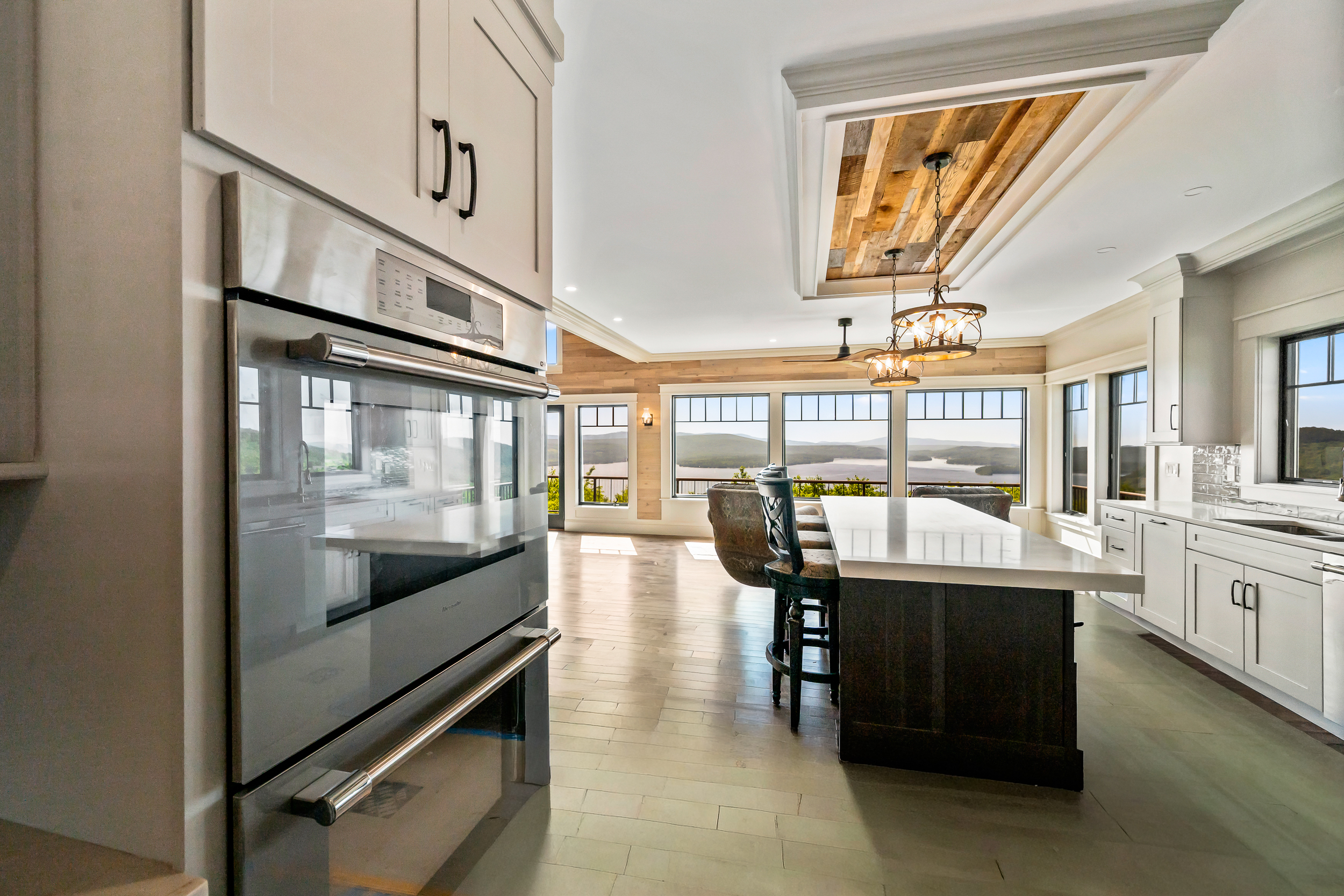 This stunning open-concept kitchen features elegant white shaker-style cabinetry, a contrasting dark island with a crisp white countertop, and high-end stainless steel appliances including a built-in double wall oven. The space is anchored by a coffered ceiling with reclaimed wood inlay and sophisticated pendant lighting, all while maximizing the breathtaking panoramic views of the water and mountains through a expansive wall of windows.