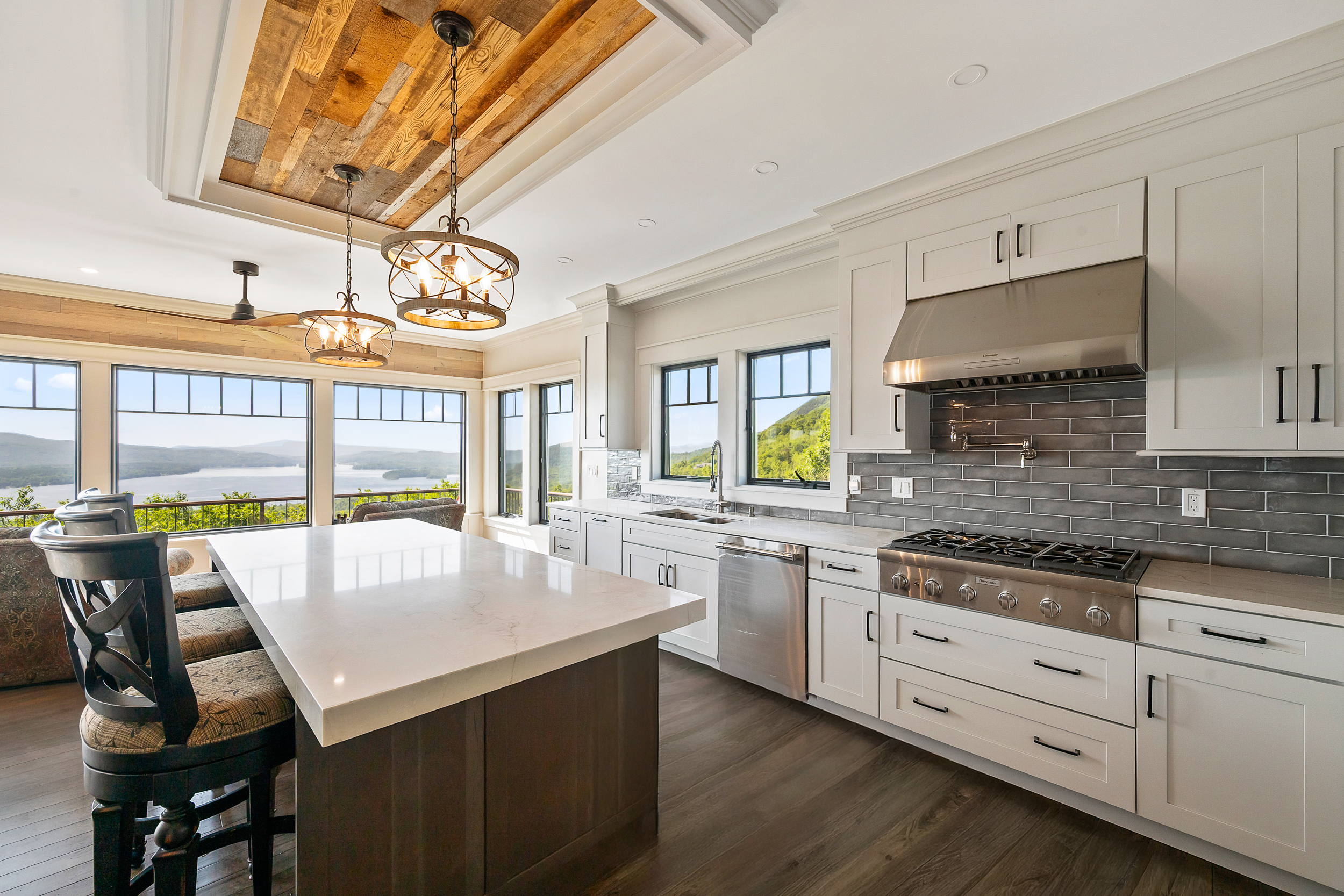 This bright, upscale kitchen features a clean aesthetic with white shaker-style cabinetry, quartz countertops, and a large center island with wooden detailing. Notable features include high-end stainless steel appliances, a stylish gray subway tile backsplash, and unique rustic wood plank ceiling accents. Large windows provide stunning, unobstructed panoramic views of a lake and mountainous landscape, perfectly blending indoor luxury with the natural surroundings.