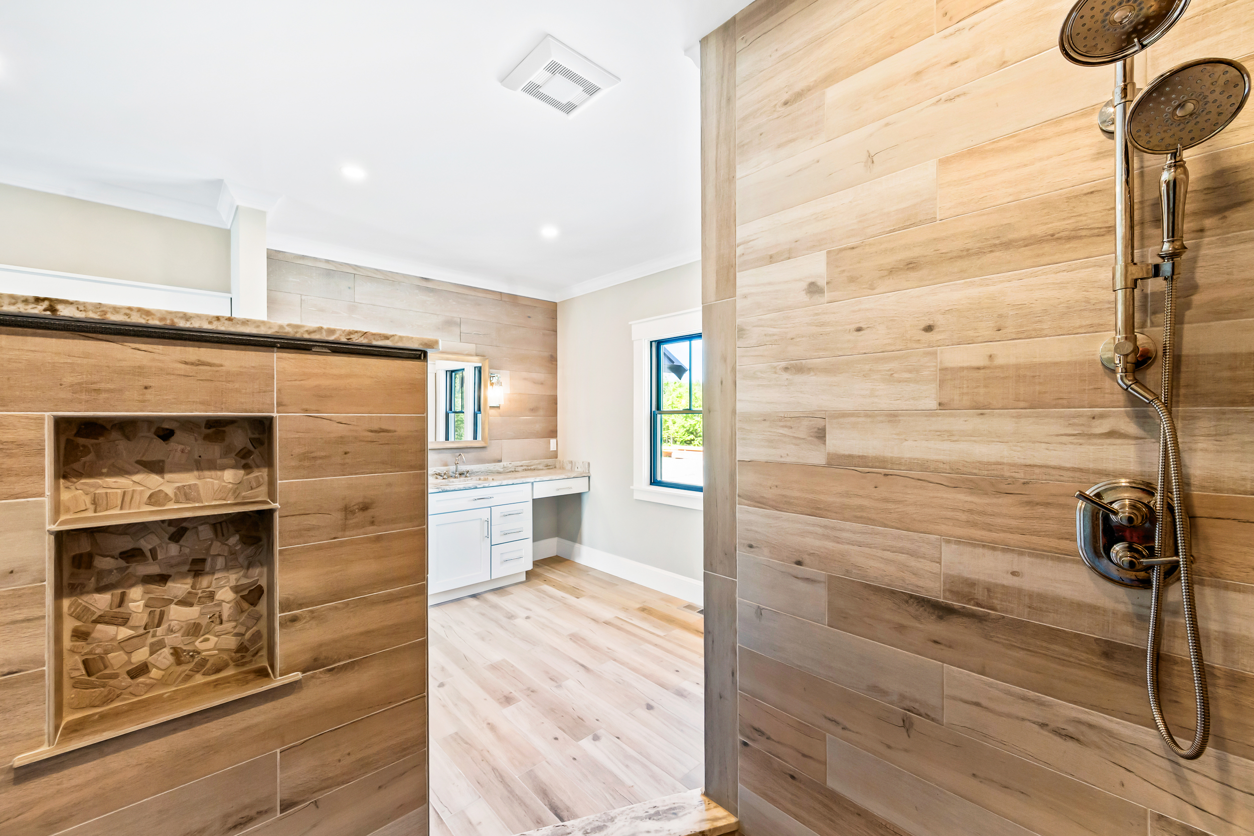 This modern bathroom features a walk-in shower area clad in large-format wood-look tile with a built-in recessed niche for storage. The space seamlessly transitions into a bright vanity area, complete with a white cabinet, stone countertop, and a clean, contemporary aesthetic. High-quality fixtures and natural wood textures create a luxurious, spa-like atmosphere throughout the room.