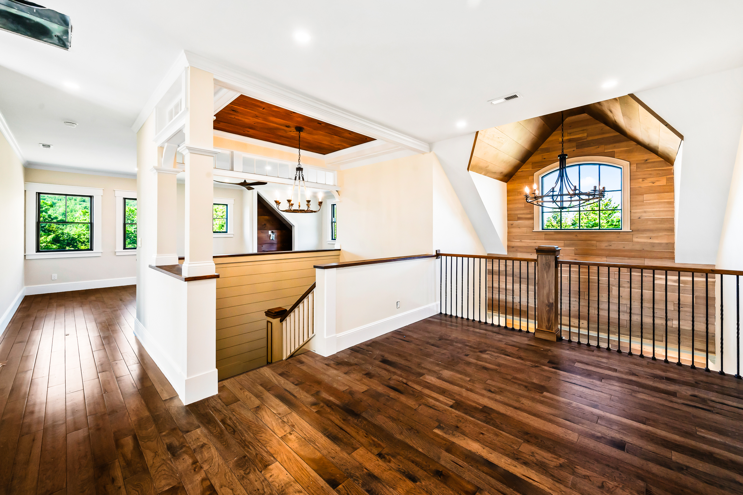 This bright and airy landing features rich hardwood flooring and a sophisticated blend of white trim with warm wood accents, including a stunning plank-ceilinged alcove. A handsome staircase with a white railing leads downward, complemented by an impressive vaulted ceiling and iron-style chandelier that highlights the architectural beauty. The space feels open and inviting, perfect for transition areas in a modern farmhouse or transitional-style home.