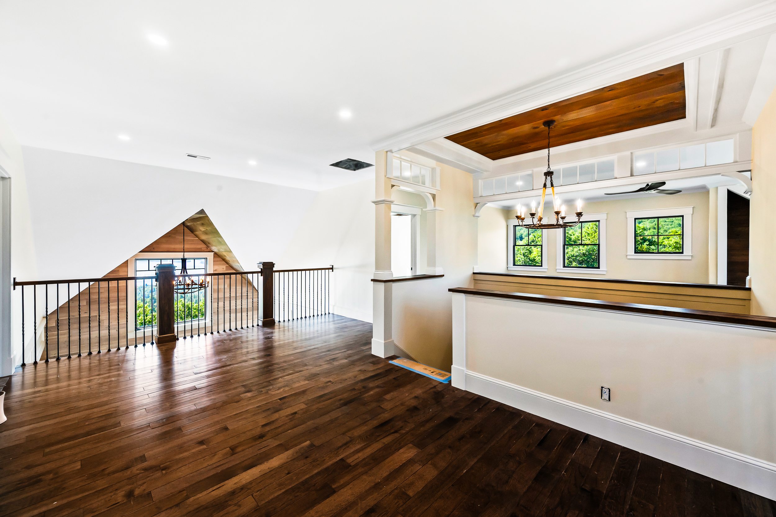 This spacious interior landing area features rich dark hardwood floors, a dramatic A-frame ceiling detail with wooden paneling, and an elegant staircase transition. Architectural elements include a decorative chandelier, expansive windows offering abundant natural light and scenic views, and high-end millwork with custom stair railings. The space feels open, airy, and sophisticated, making it a well-designed highlight of the home's upper level.