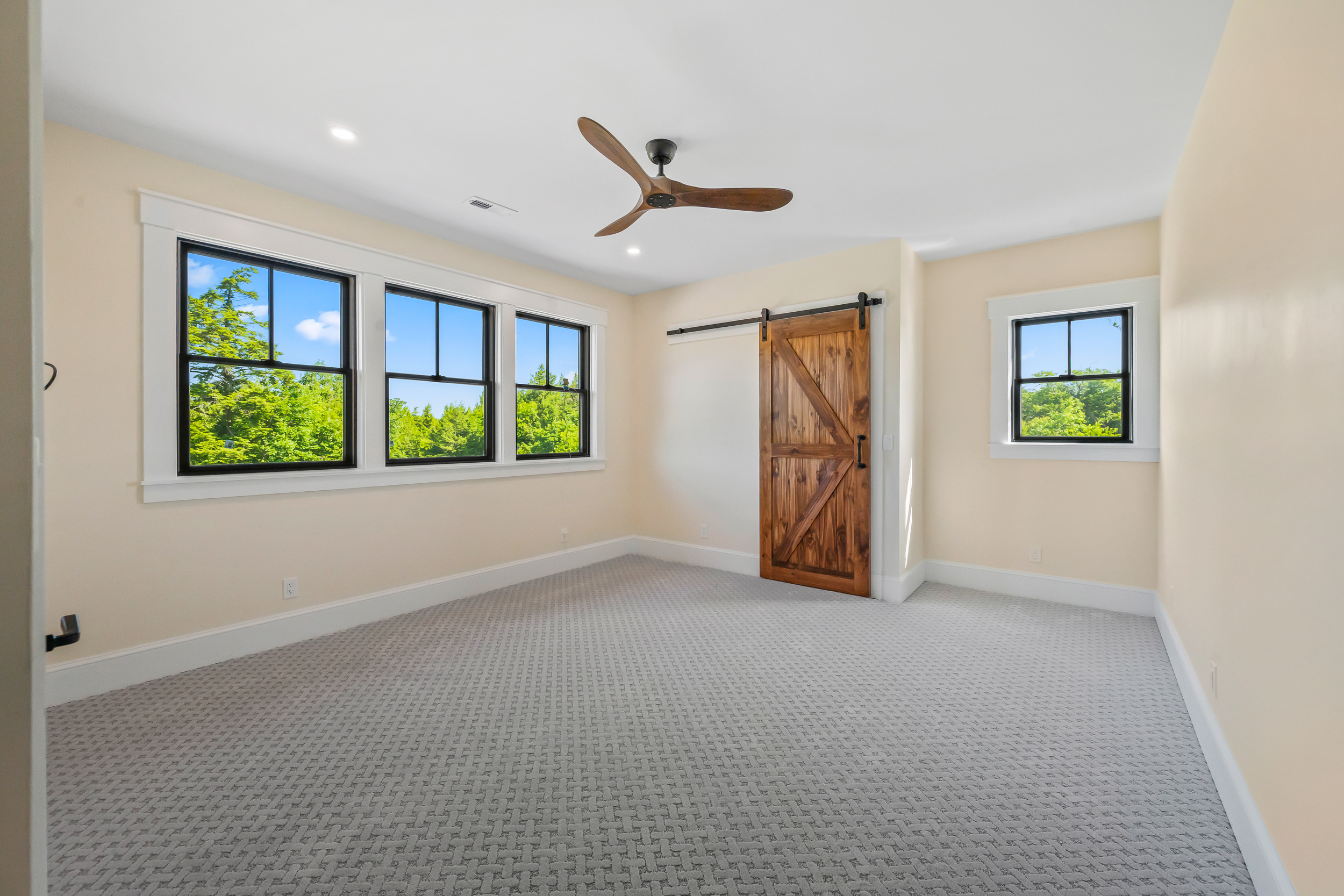 This spacious, light-filled bedroom features neutral walls, patterned wall-to-wall carpeting, and a unique wooden barn door. Large, multi-pane windows offer views of the surrounding trees, while a sleek ceiling fan adds a modern touch. The room provides a clean, versatile layout perfect for a guest bedroom or additional living space.
