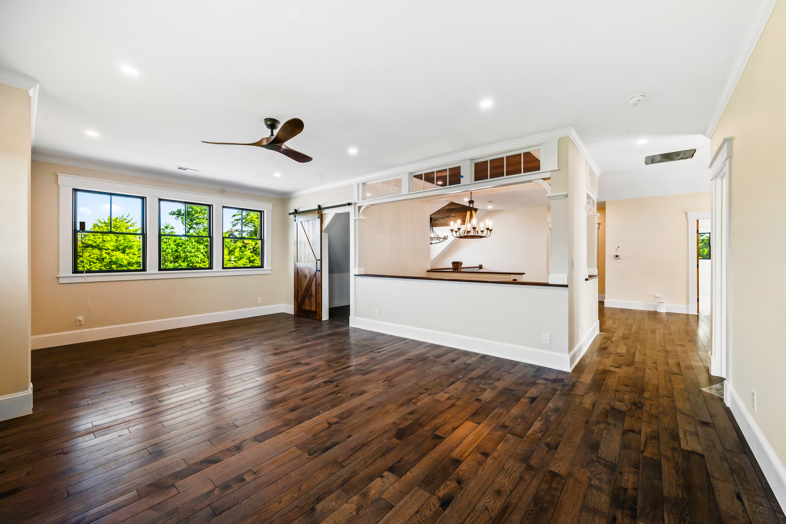 This spacious, open-concept living area features rich, dark-stained hardwood flooring and neutral paint tones, creating a welcoming and versatile atmosphere. The room is highlighted by a unique half-wall partition with transom-style windows and a rustic sliding barn door, adding architectural interest and character. Natural light pours in through the large windows, which provide a lush, green view of the outdoors.