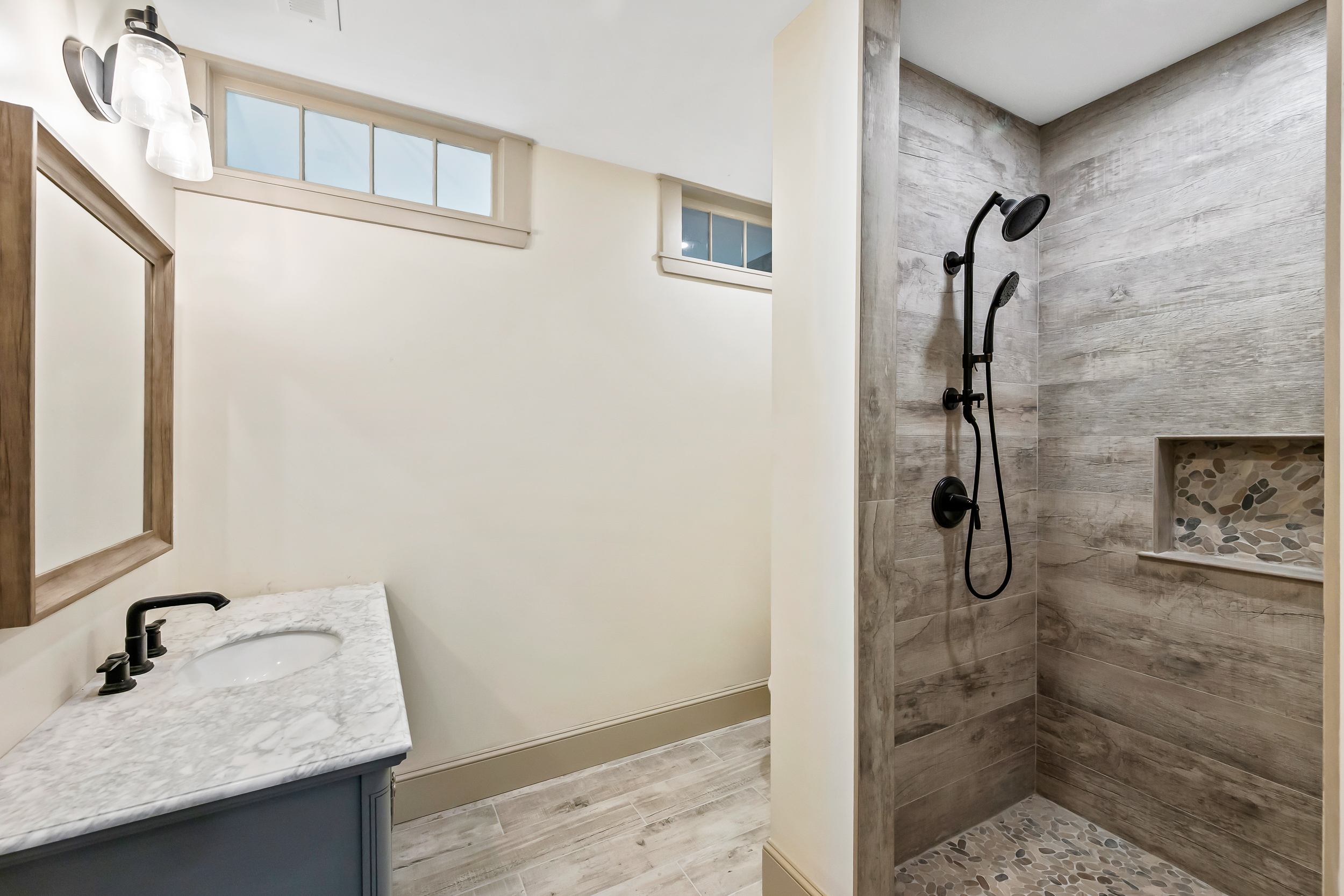 This contemporary bathroom features a sophisticated design with a marble-topped vanity and a walk-in shower encased in wood-look tile. The space includes natural elements like a stone tile shower floor and a pebble-inlaid shower niche, complemented by modern matte black fixtures and clean lines. The overall impression is one of stylish, rustic-modern comfort.