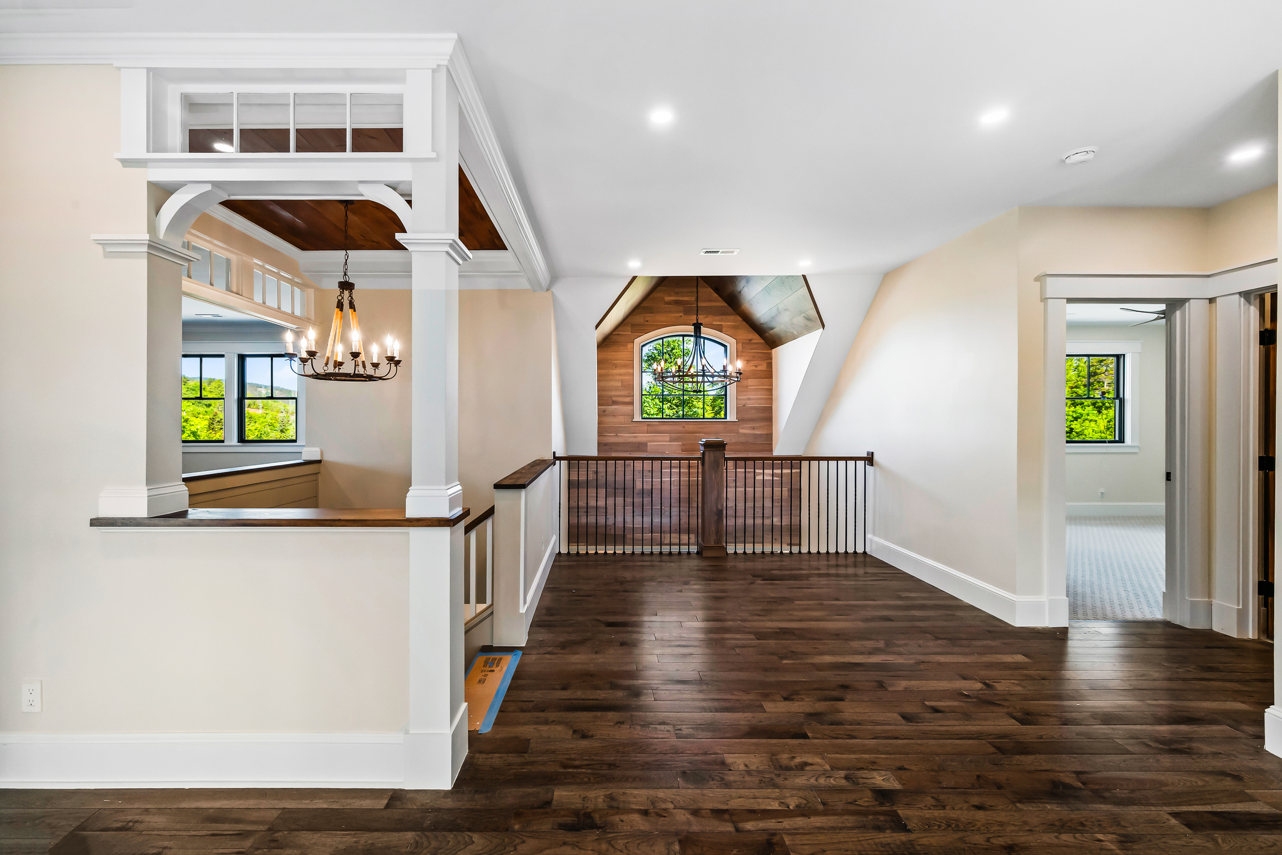 This spacious, light-filled landing area showcases a sophisticated blend of traditional craftsmanship and farmhouse-inspired design. Notable features include wide-plank hardwood flooring, ornate custom woodwork around the staircase, and a striking wooden accent wall surrounding a vaulted window. The space is anchored by two elegant chandeliers, creating a grand and inviting transitional area that flows into other parts of the home.