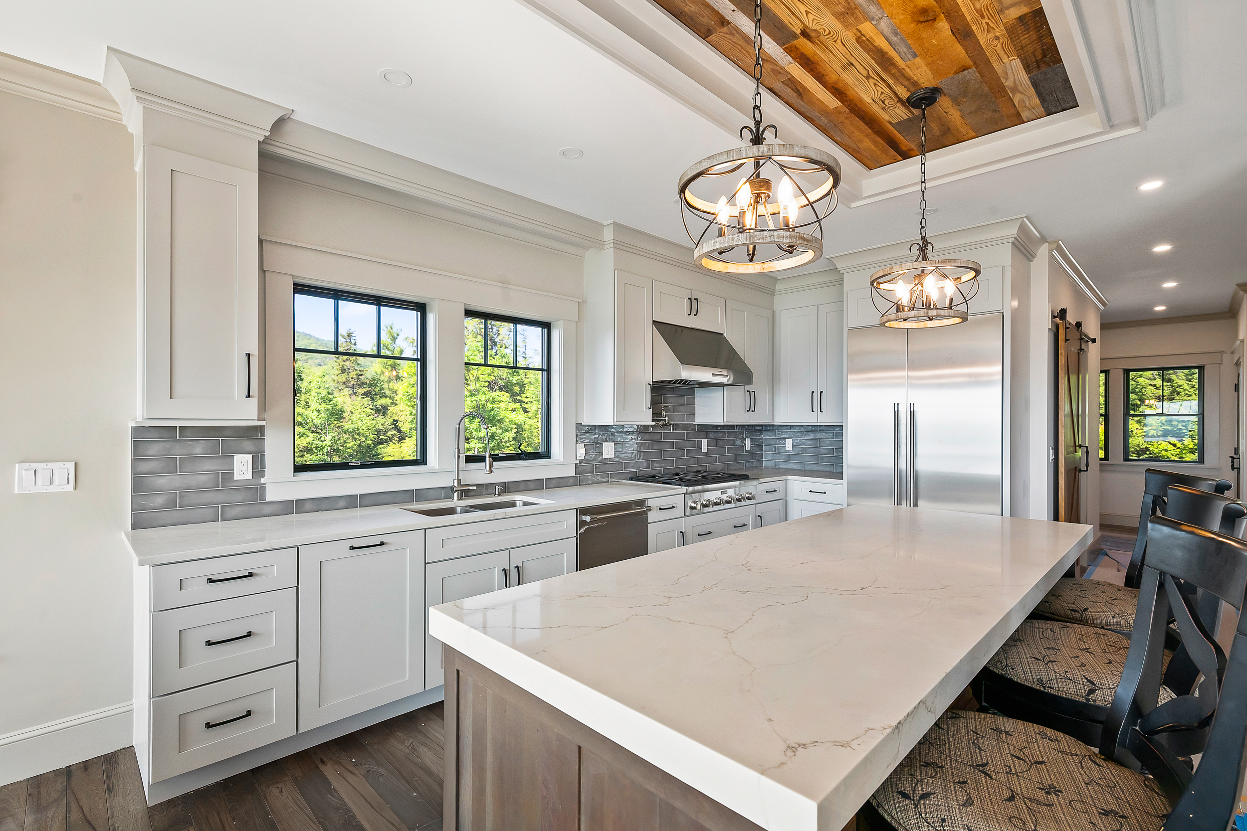 This modern kitchen features crisp white cabinetry contrasted by a dark wood island, stainless steel appliances, and elegant gray subway tile backsplash. The space is highlighted by a stunning wooden slat ceiling detail and two rustic-industrial style orb pendant lights hanging over the large white quartz-topped island. Large windows offer plenty of natural light and scenic woodland views, creating an inviting and sophisticated aesthetic.