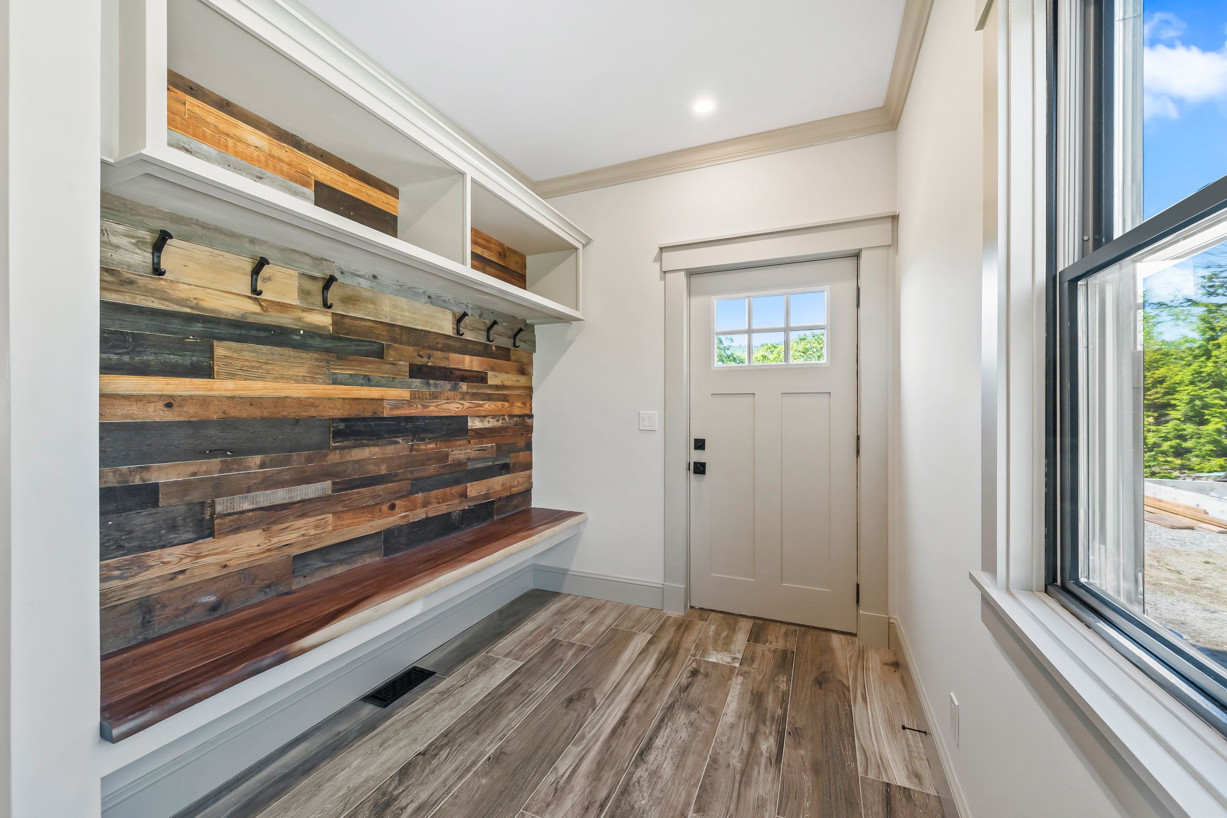 This modern mudroom space features a distinct plank-style reclaimed wood accent wall complete with sturdy coat hooks and overhead storage cubbies. A rich wooden bench provides functional seating, while light-toned, wood-look flooring and a bright entryway door create a welcoming, farmhouse-inspired transition area. Large windows invite natural light, highlighting the clean white trim and thoughtful architectural character of the entry.