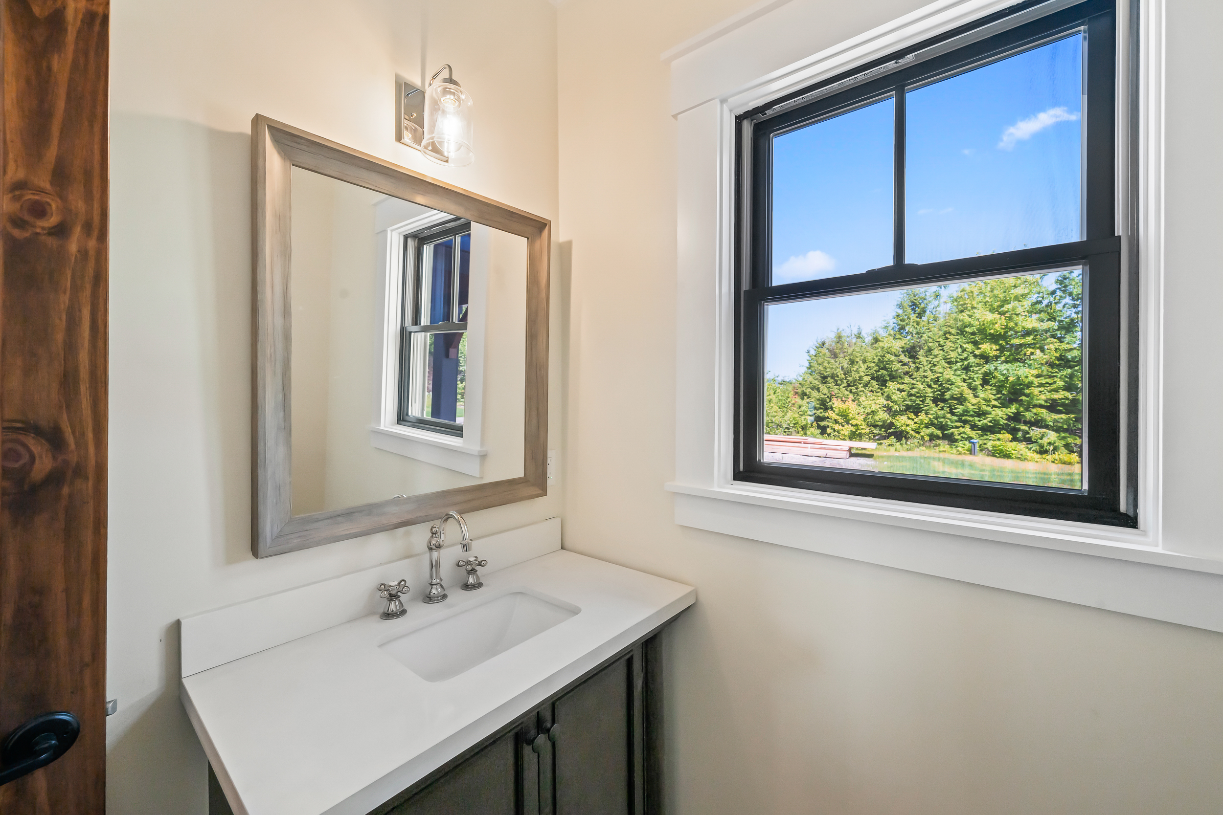 This bright and inviting bathroom vanity area features a single sink with a polished chrome faucet set into a clean, white countertop. A handsome wood-framed mirror hangs above, reflecting the natural light pouring in from the adjacent window, which offers a serene view of the surrounding greenery. The dark-toned vanity cabinet provides a sophisticated contrast to the light neutral walls, creating a polished and modern aesthetic.