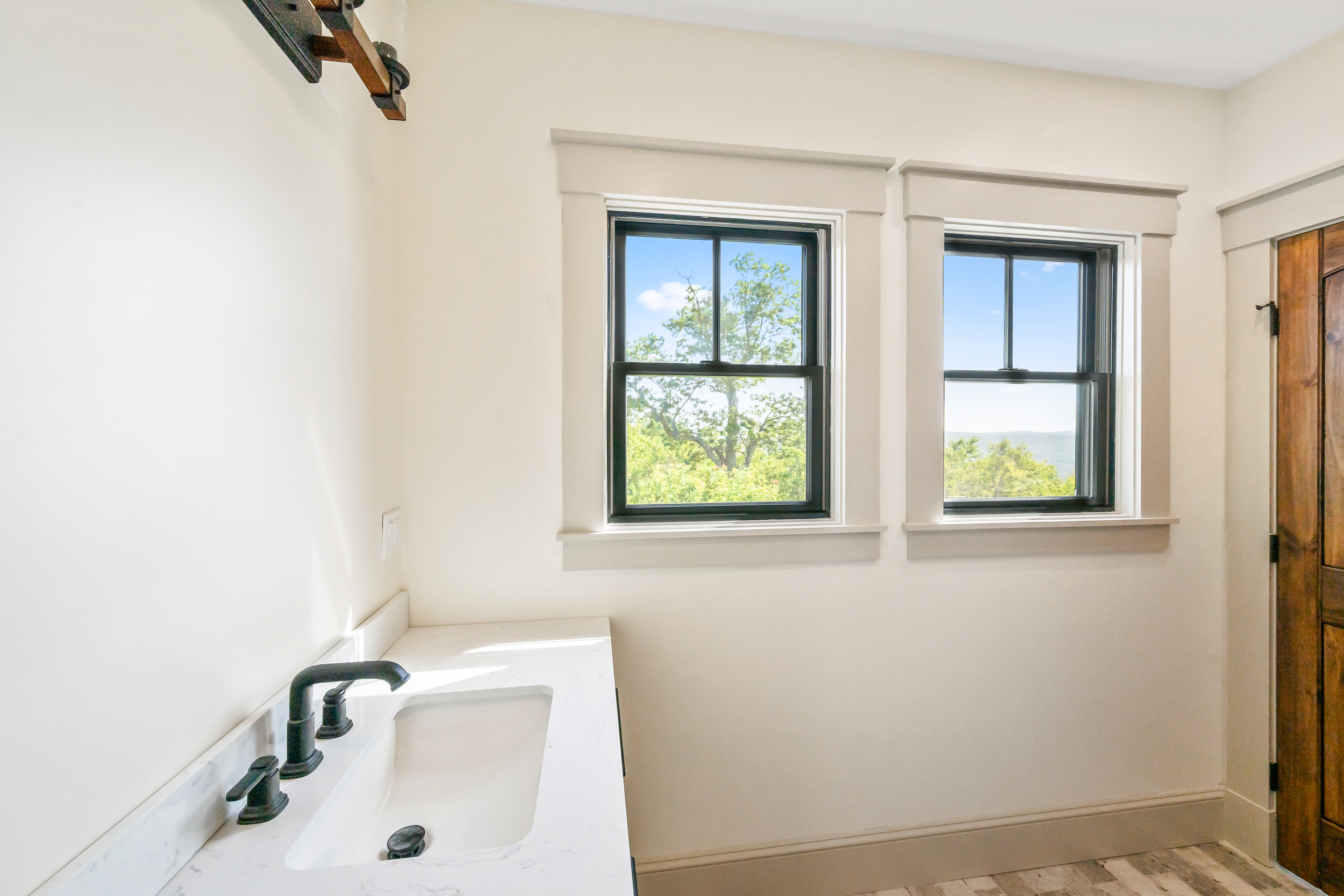 This bathroom features a sleek vanity with a white marble countertop and a modern dark-finished faucet. Two large windows with dark frames provide natural light and expansive views of the surrounding trees and landscape. The neutral, minimalist design is accented by warm wooden door trim and high-quality finishes, creating a bright and airy atmosphere.