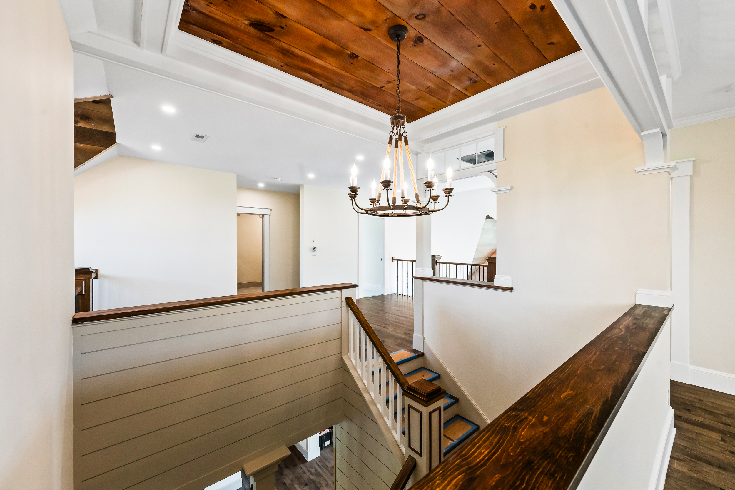This interior view showcases an elegant landing area featuring a warm, stained-wood plank ceiling and an impressive, rustic-style chandelier. The space flows into a staircase with classic white railings and stained-wood handrails, highlighting a blend of modern farmhouse design and traditional architectural elements. Bright, neutral wall tones combine with rich wood textures to create an open and inviting atmosphere at the top of the stairs.