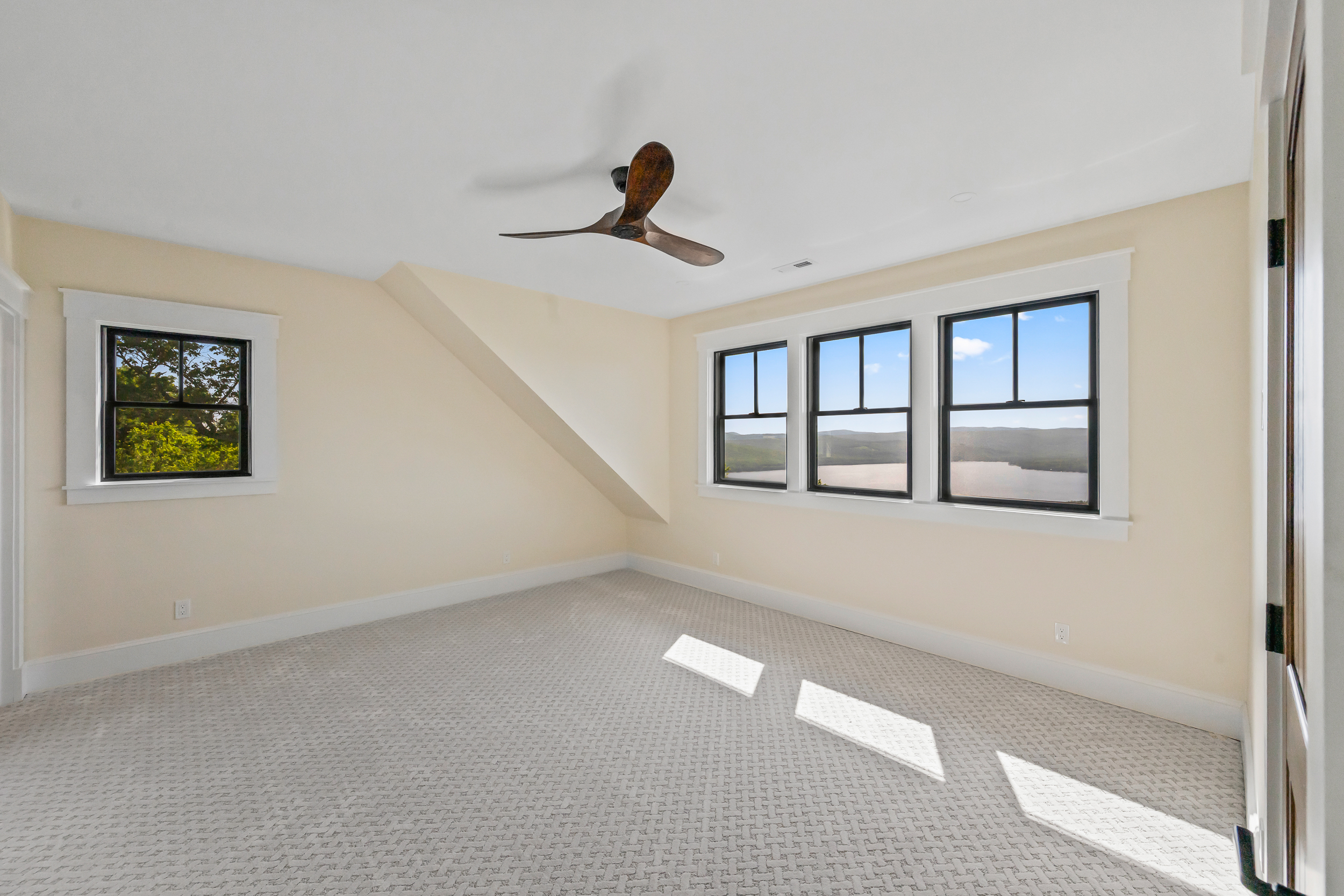 A bright and neutrally toned bedroom featuring a clean, minimalist aesthetic with cream-colored walls and patterned carpeting. An angled ceiling adds architectural interest, while large windows offer a scenic, panoramic view of the nearby water and landscape. The room is further highlighted by a stylish wood-finish ceiling fan, creating a welcoming and peaceful atmosphere.