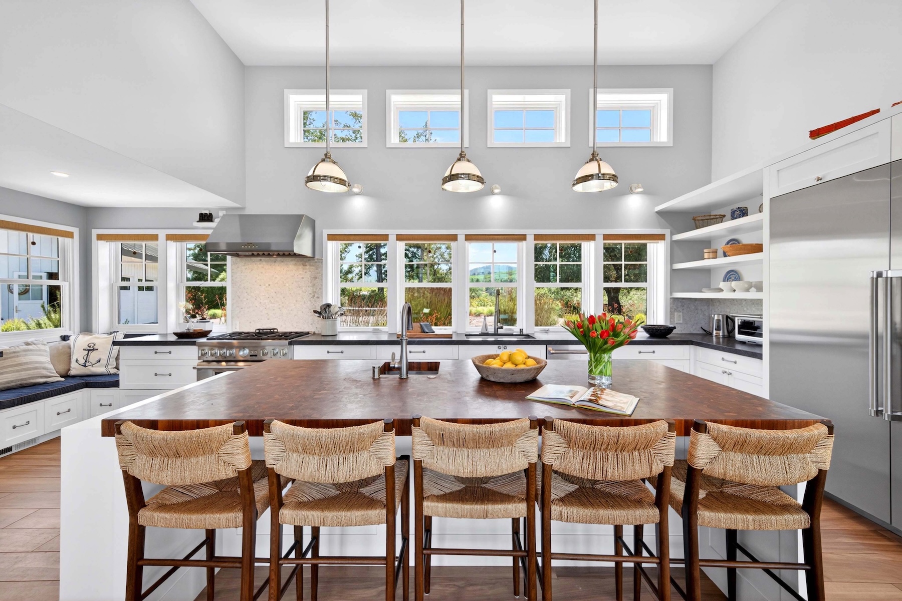 This bright and airy kitchen features a large center island with a warm wood countertop and five woven stool seats, making it an excellent space for entertaining. The white cabinetry and neutral color palette are complemented by a stainless steel stove, modern pendant lighting, and abundant natural light from the large windows and transom windows above. A cozy window seat in the corner adds charm, while open shelving provides practical storage, resulting in a clean, farmhouse-inspired aesthetic.