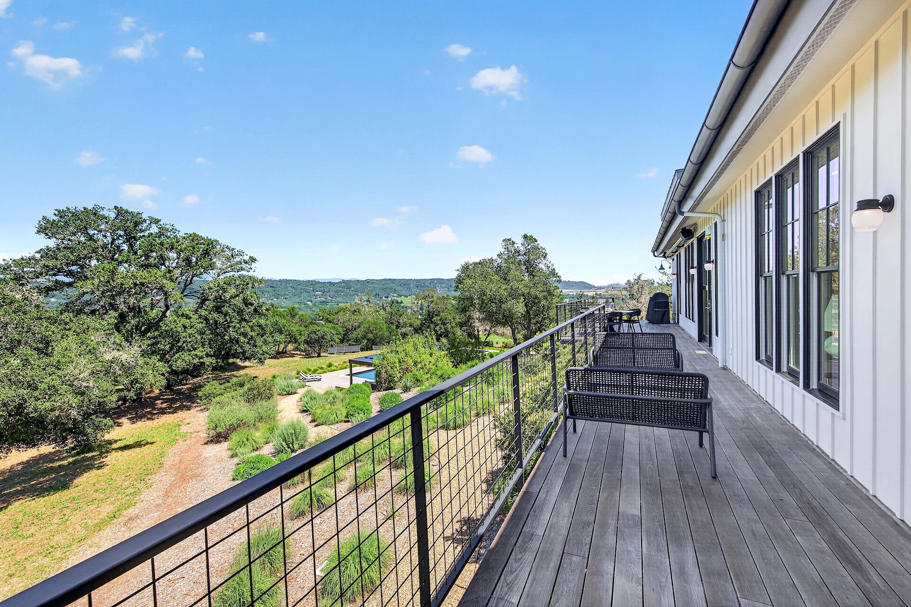 This spacious, modern balcony features wooden decking and a sleek black metal mesh railing that provides unobstructed views of the surrounding landscape. The outdoor area is furnished with contemporary black seating, creating an inviting space to enjoy the elevated scenery overlooking the yard, greenery, and distant hills. The home's white board-and-batten siding and exterior sconce lighting lend a stylish, farmhouse-inspired aesthetic to the outdoor living area.