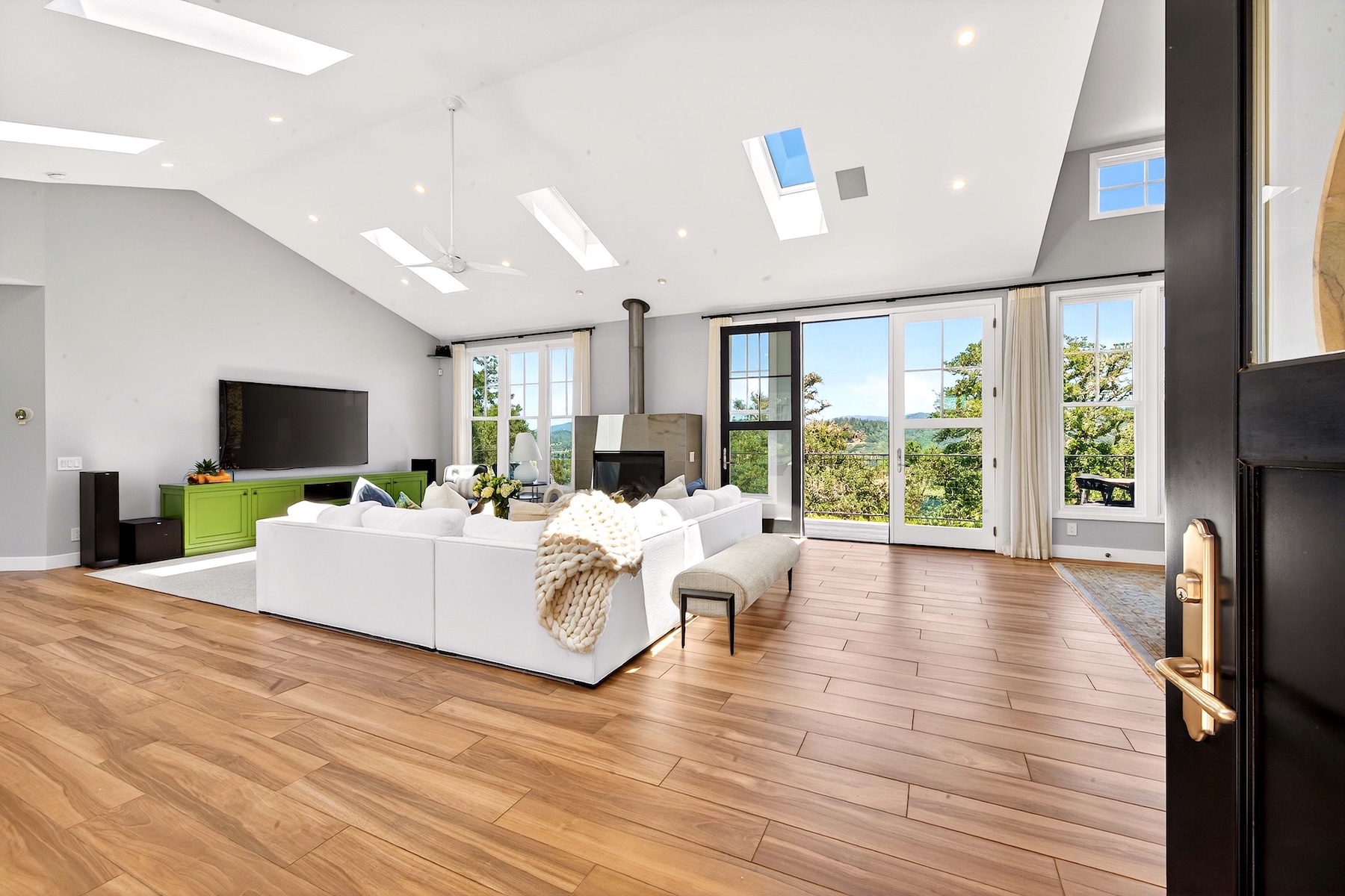 This spacious and airy living room features a vaulted ceiling with multiple skylights and stunning hardwood flooring throughout. The room is anchored by a large, modern white sectional sofa and a sleek fireplace, while the expansive glass doors offer a seamless transition to the outdoors with scenic foliage views. A bright green media cabinet adds a pop of color to the otherwise neutral, contemporary aesthetic.