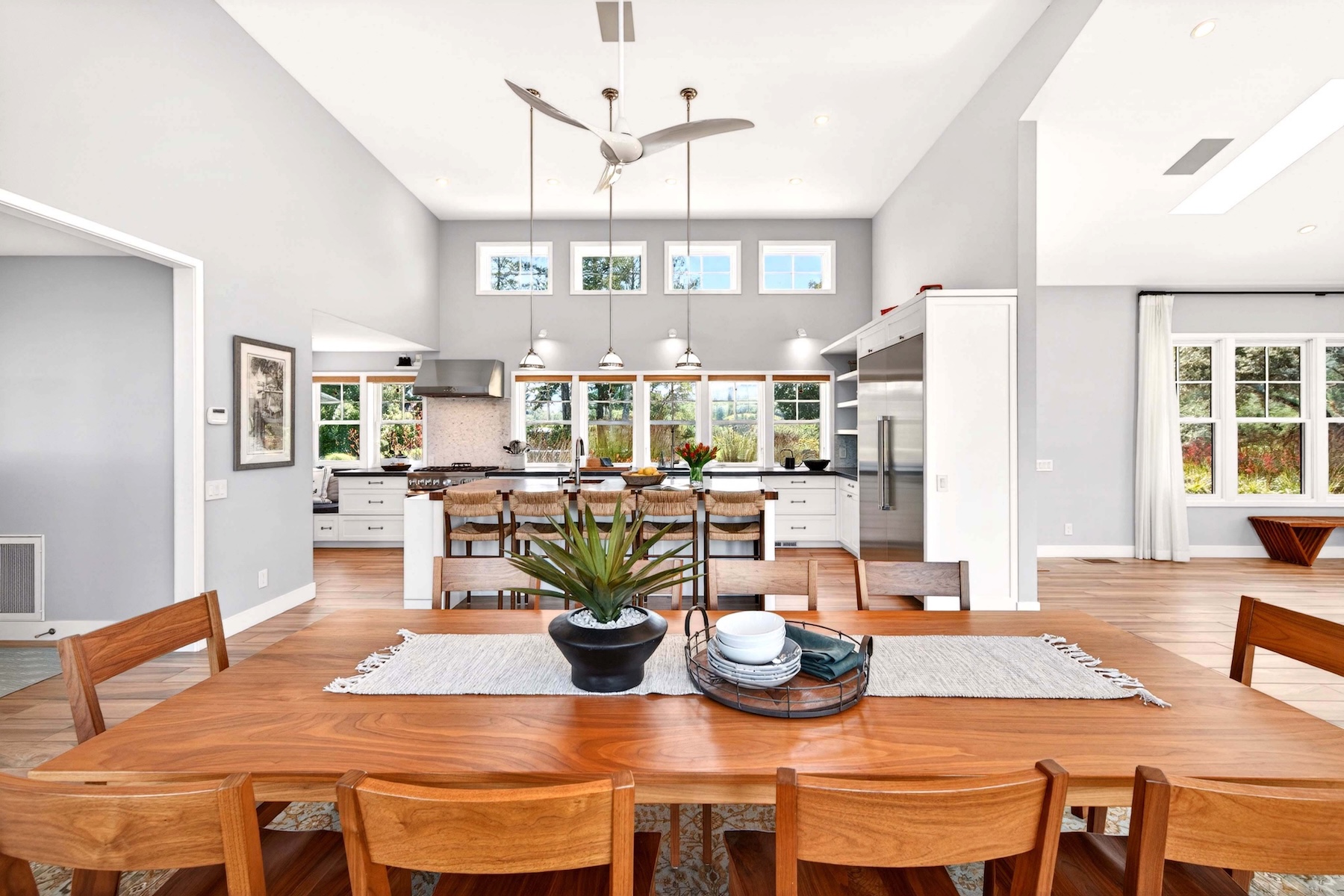 This bright, open-concept dining area features a large natural wood table as the central focal point, leading directly into a modern, airy kitchen. The seamless flow between the dining space and kitchen, combined with high ceilings and pendant lighting, creates an inviting atmosphere perfect for entertaining. Large windows throughout the space flood the room with natural light, highlighting the well-maintained interior design.