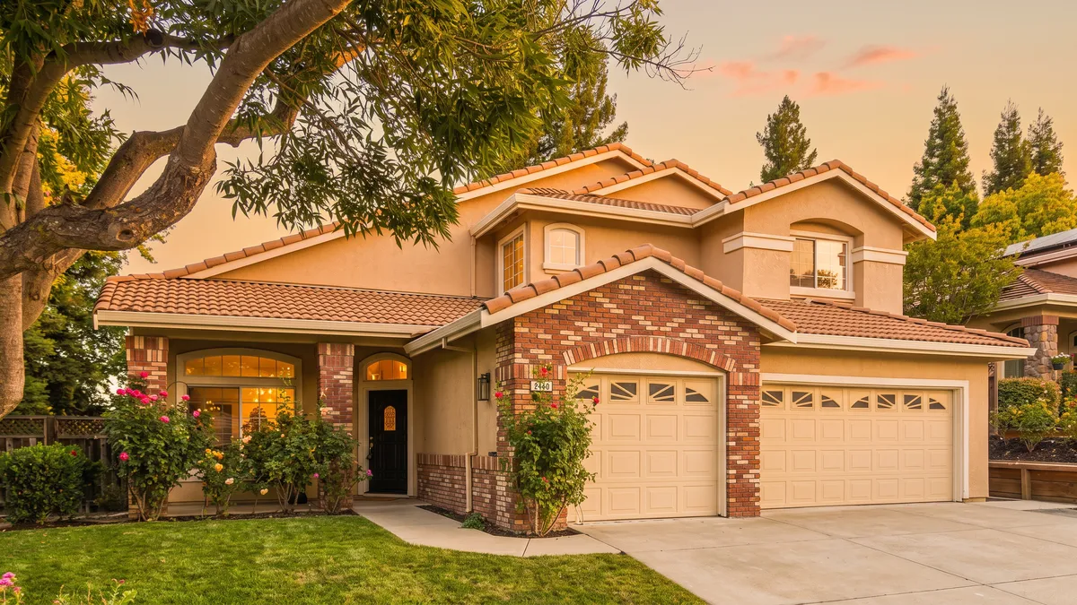 This charming two-story suburban home features a traditional architectural style with a mix of tan stucco and red brick facade accents. The property includes a spacious three-car garage, an inviting front entryway, and well-maintained landscaping with flowering bushes. Captured during a warm sunset, the home exudes a welcoming and well-cared-for appeal, perfect for a family residence.