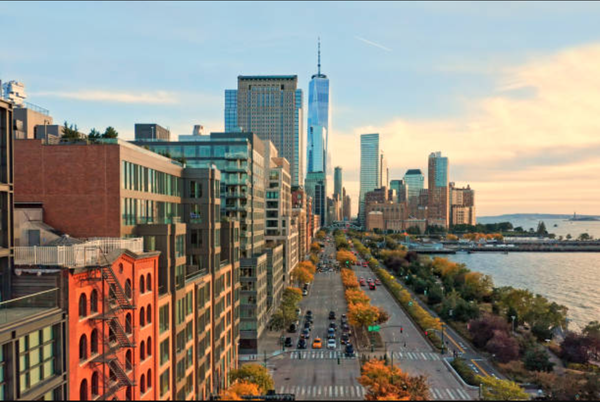 This photo showcases an expansive aerial view of New York City, emphasizing major landmarks like the One World Trade Center against a backdrop of the skyline and waterfront. The image highlights the urban landscape, with streets lined with trees in autumnal colors and bustling with traffic. It would serve to illustrate the accessibility and desirability of the location within the city.
