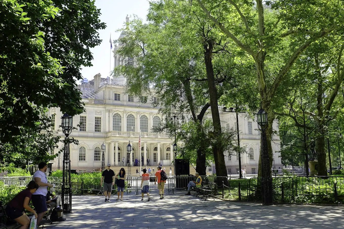 This image showcases the front of a grand, white multi-story building, likely a city hall or similar municipal building, framed by lush green trees and a well-maintained plaza. People are seen walking and relaxing in the foreground, adding a sense of activity and community. The overall impression is one of historical significance and civic pride, suggesting a prime location in a vibrant urban center.