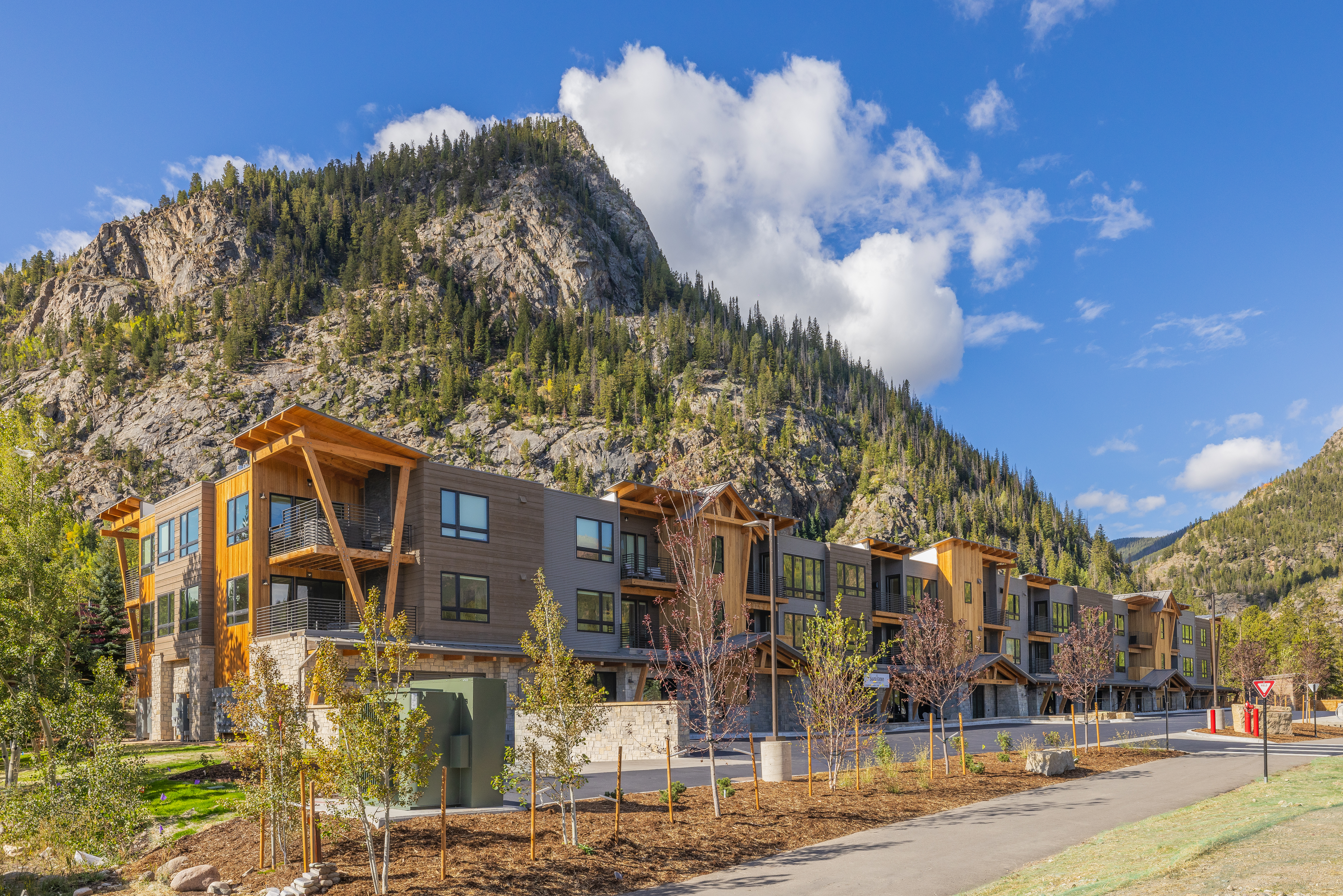 This image showcases the front exterior of a modern multi-unit building nestled against a mountain backdrop. The building features a combination of wood and gray siding, with visible balconies. Landscaping includes young trees and a well-maintained walkway, enhancing the property's curb appeal.
