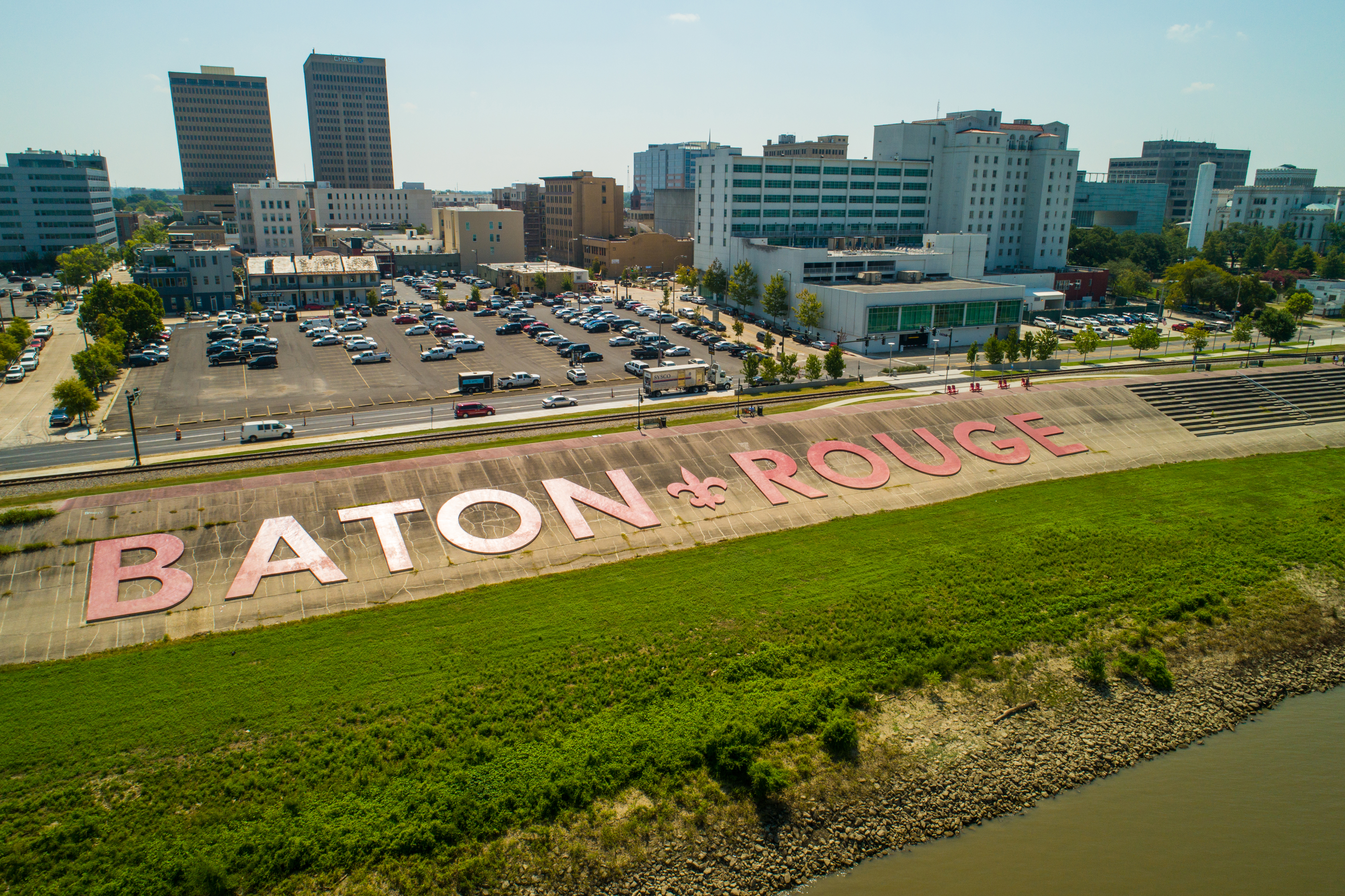 This aerial view captures the prominent downtown Baton Rouge riverfront, featuring the iconic 'BATON ROUGE' lettering prominently displayed on the levee wall. The scene includes an expansive city skyline with mid-to-high-rise buildings, a large public parking lot, and the adjacent Mississippi River shoreline. The perspective highlights the city's urban landscape and its connection to the river, making it an excellent representation of the downtown area.