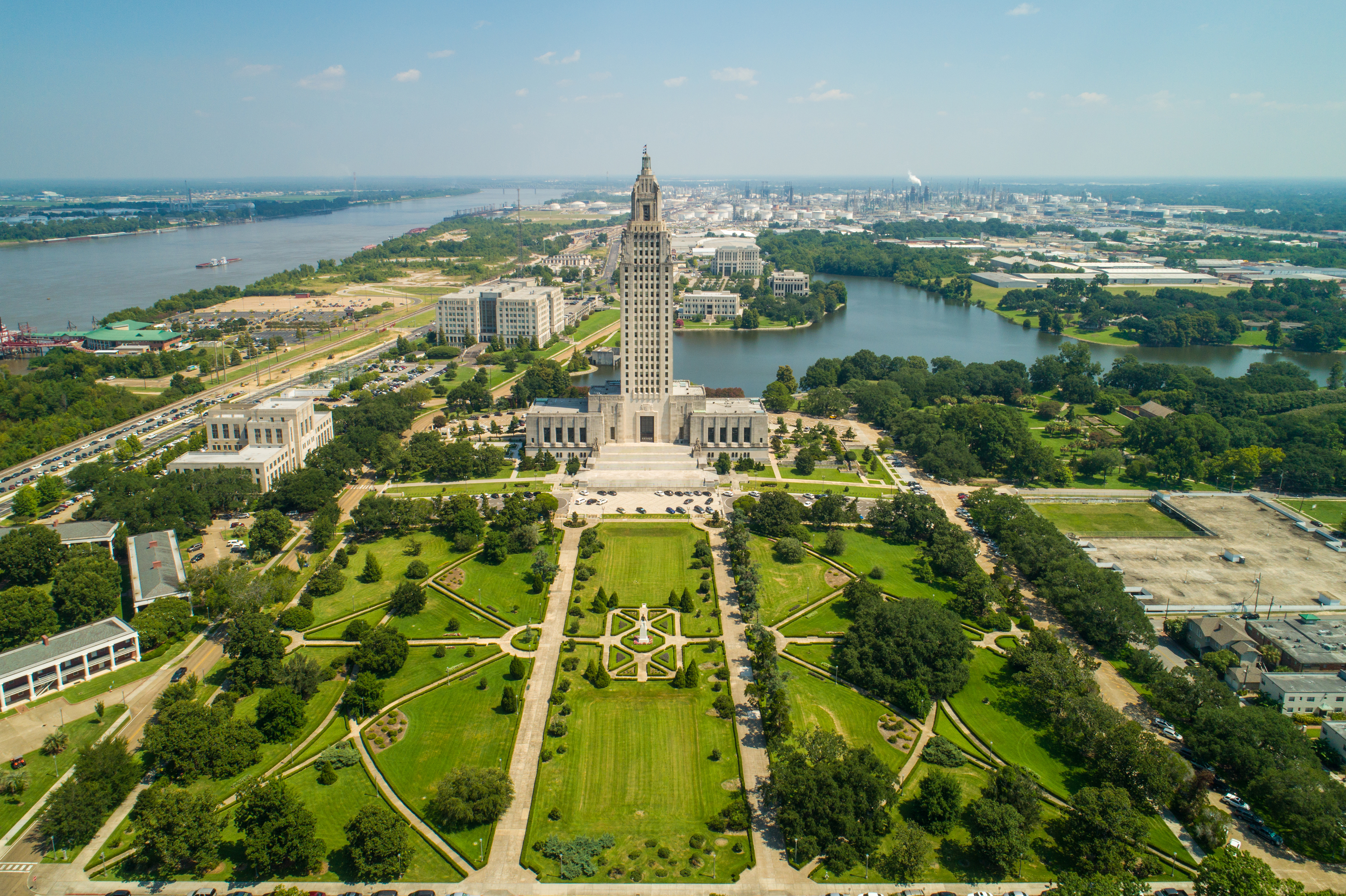 This stunning aerial view captures the Louisiana State Capitol building as the centerpiece of a meticulously landscaped government complex. The grand architectural structure is framed by expansive, symmetrical gardens and green space, leading to a scenic riverfront setting. The combination of historic monumental design and lush, well-maintained park grounds creates a majestic and impressive impression.
