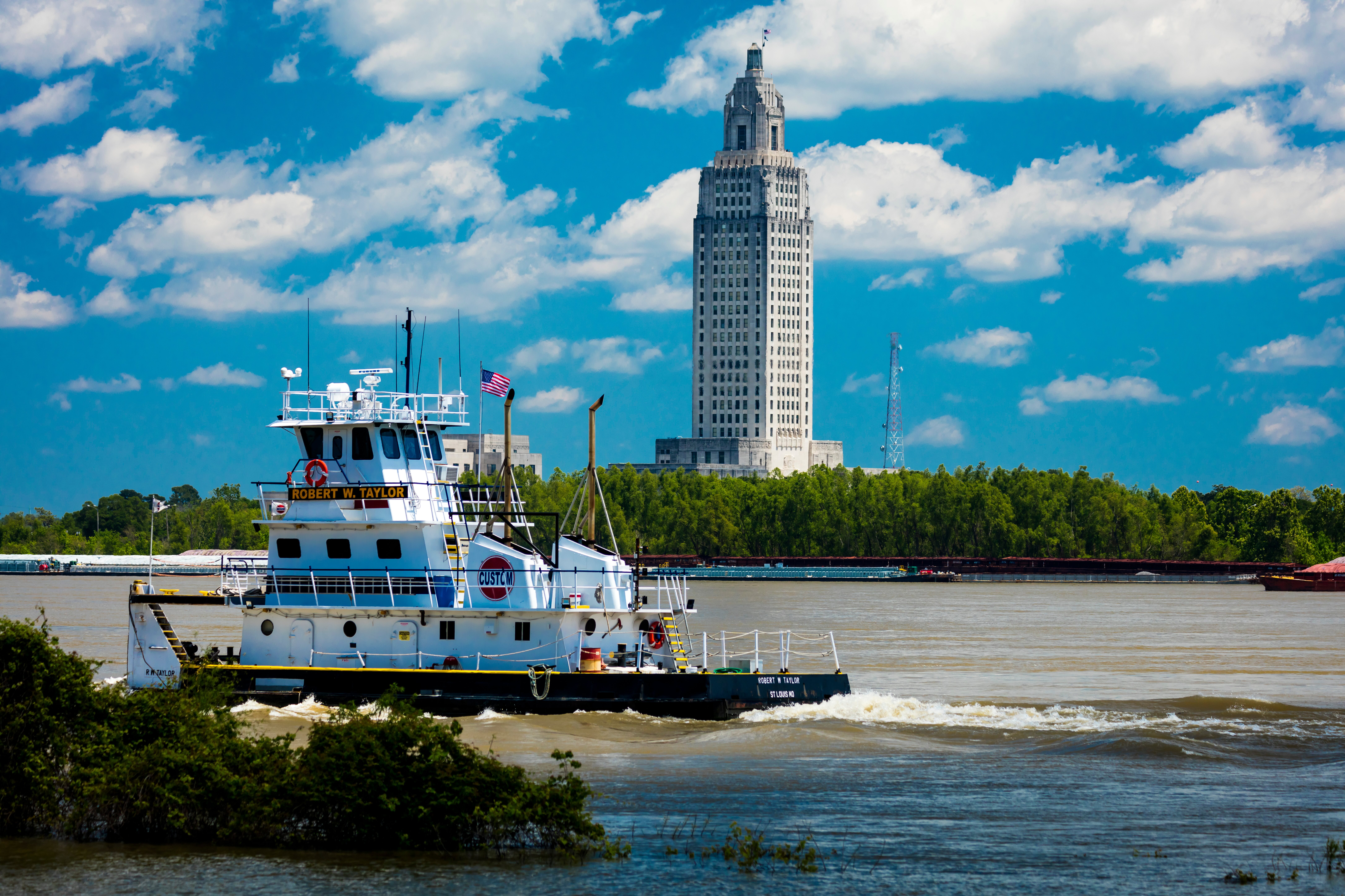This image captures a tugboat, named 'Robert W. Taylor', navigating a river with the Louisiana State Capitol building in the background. The scene highlights the bustling industrial activity along the riverfront, offering a scenic view of a major landmark. The bright blue sky with scattered clouds provides a picturesque backdrop for the urban and maritime setting.