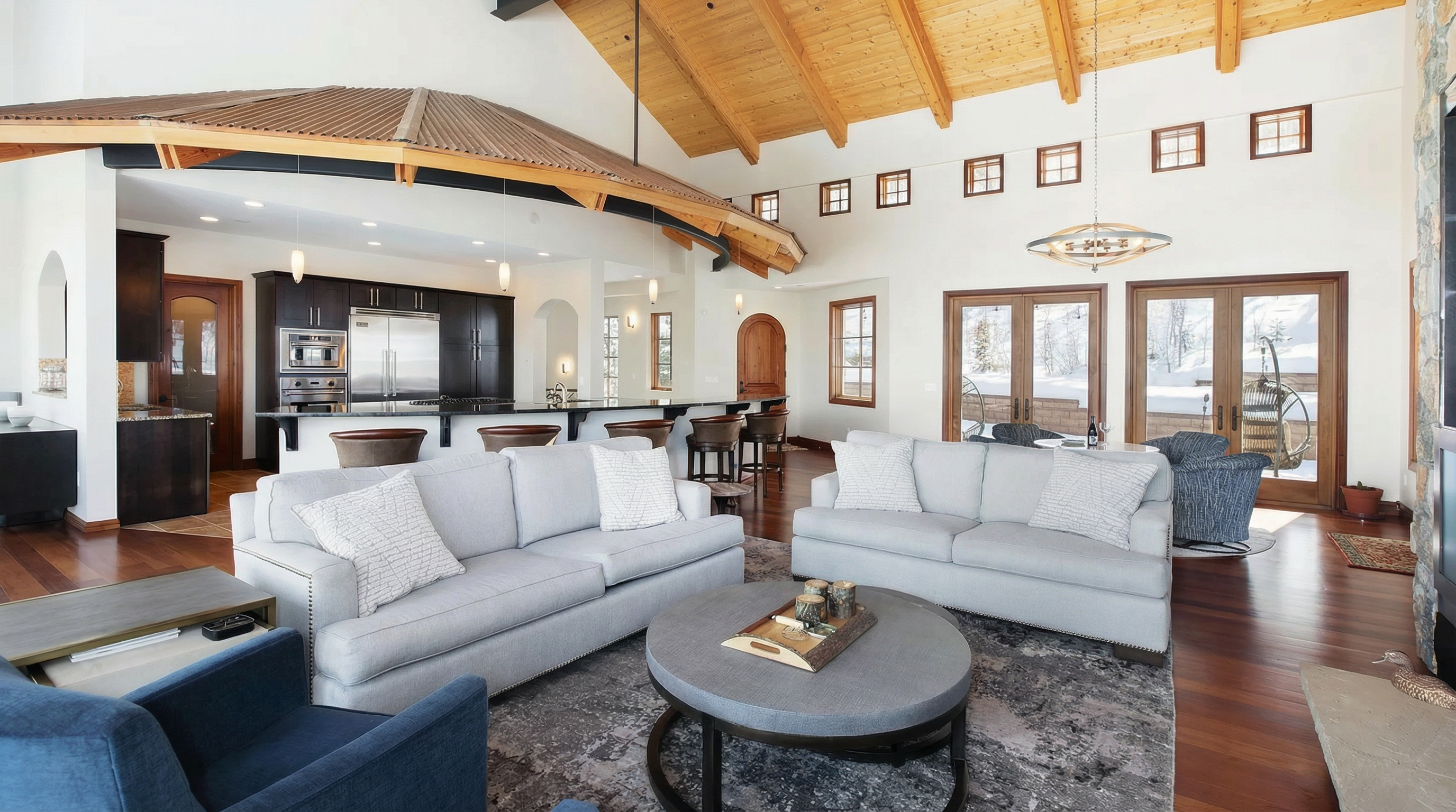This is a well-appointed living room featuring two light gray sofas, a textured gray rug, and a round coffee table. The open floor plan seamlessly integrates with the kitchen area. The room has a high, vaulted ceiling with exposed wooden beams, creating a spacious and inviting atmosphere.