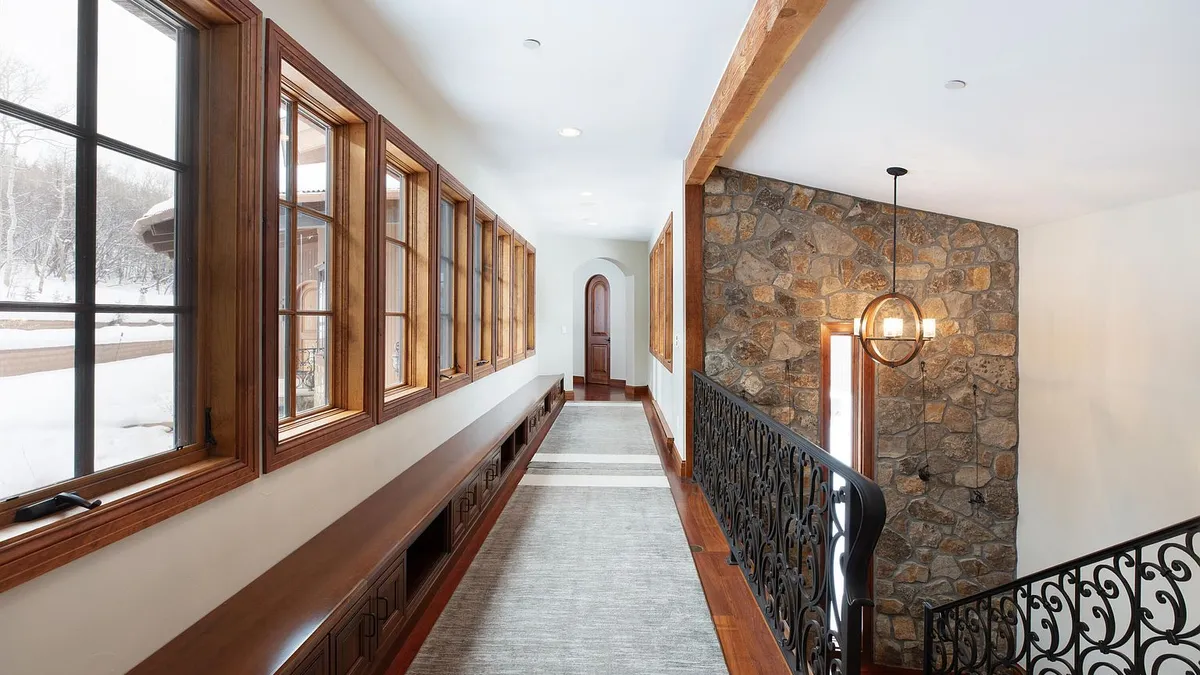 This interior shot showcases a well-lit hallway adjacent to a stairway. Natural light floods the hallway through a series of windows along one wall, complemented by recessed lighting. A decorative iron railing accents the staircase, and the stone wall adds a rustic touch to the space.
