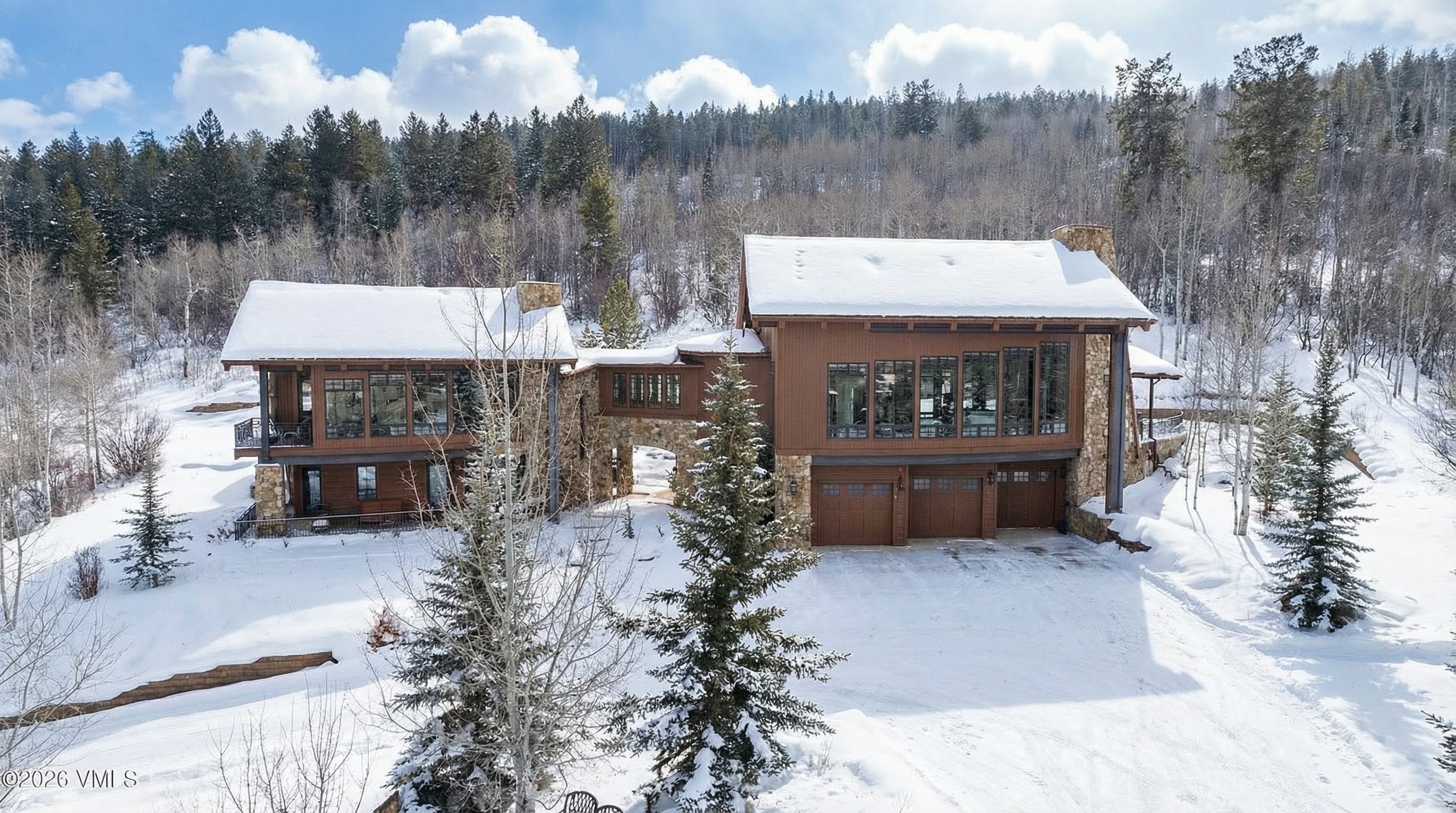 This image showcases the front exterior of a luxurious mountain residence during winter. The home features a combination of wood siding and stone accents, with snow-covered roofs and multiple windows. An arched stone walkway connects two wings of the house, leading to garage doors, set against a backdrop of snow-covered grounds and mature trees.