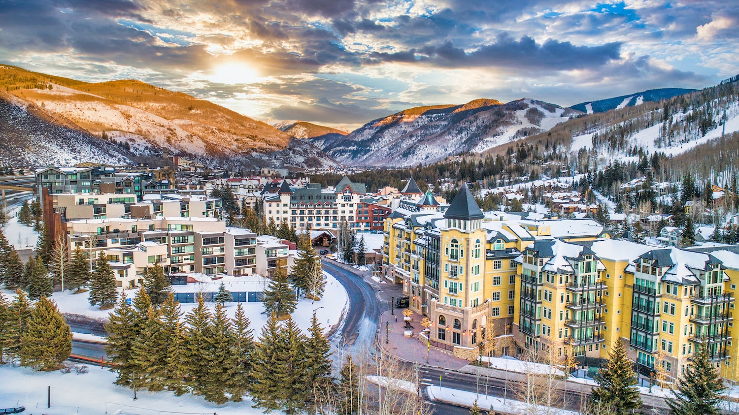 This stunning aerial view showcases a luxurious alpine resort town nestled amidst snow-capped mountains. The architecture includes a mix of modern condos and grand hotels, with meticulously maintained streets winding through the town. The image highlights the proximity to the ski slopes and the overall serene, upscale atmosphere, making it an ideal location for vacation homes or investment properties.