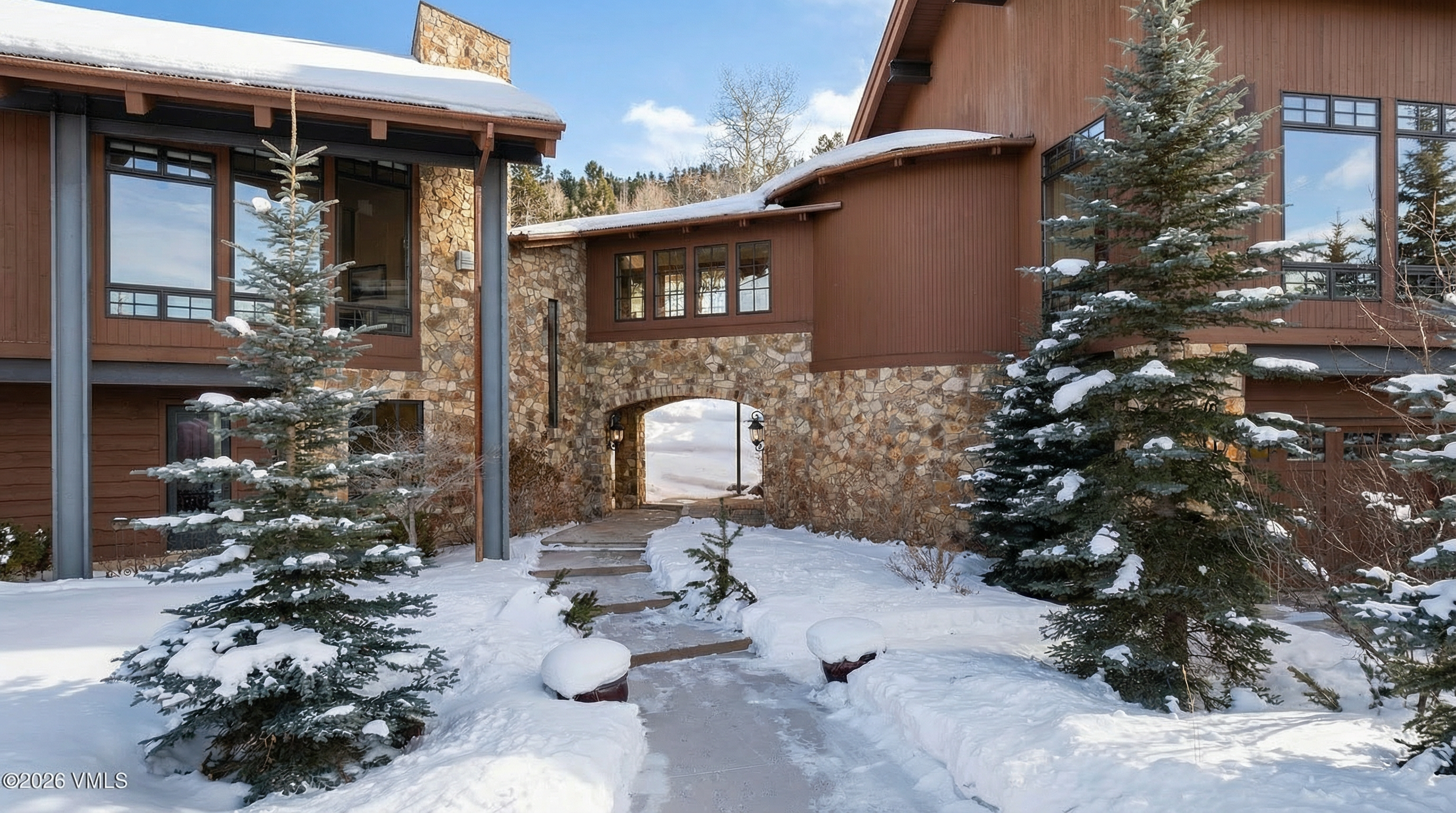 This exterior shot showcases a charming entryway of a luxury mountain home in winter. The residence features a stone archway connecting two sections of the house, with mature evergreen trees flanking the snow-covered path. The architecture combines wood and stone elements for a rustic yet refined curb appeal.