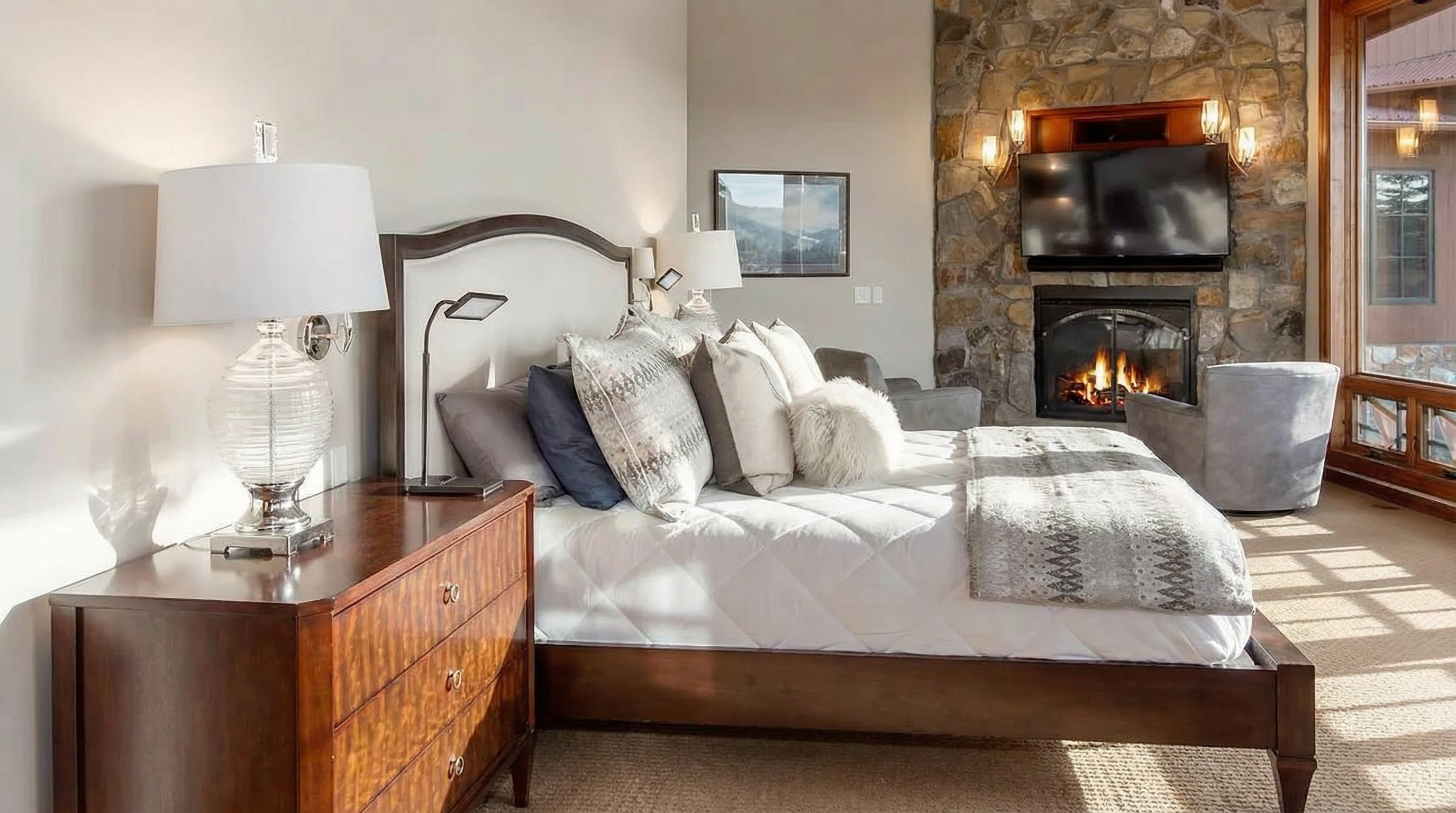 This is a luxurious primary bedroom featuring a comfortable bed with multiple pillows and a throw blanket. A stone fireplace with a mounted television creates a cozy ambiance. Natural light floods the room through a large window, complemented by elegant bedside lamps on a wooden dresser.