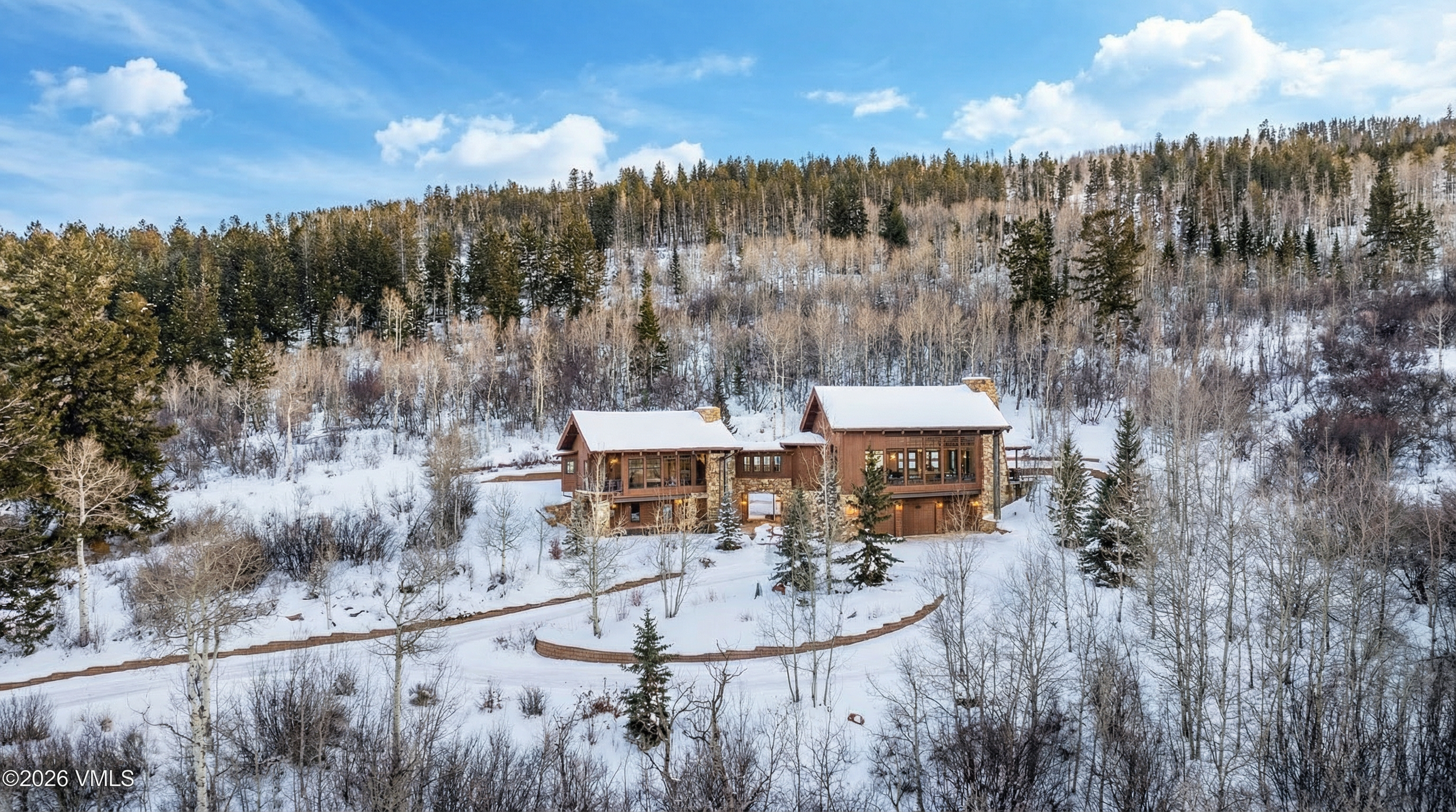 This is an exterior front view of a mountain home in a winter setting. The home is a spacious brown wood and stone house surrounded by snow-covered trees and foliage, with a winding driveway leading up to it. The house has a stone chimney and multiple windows, showcasing a rustic yet elegant design suitable for a winter retreat.