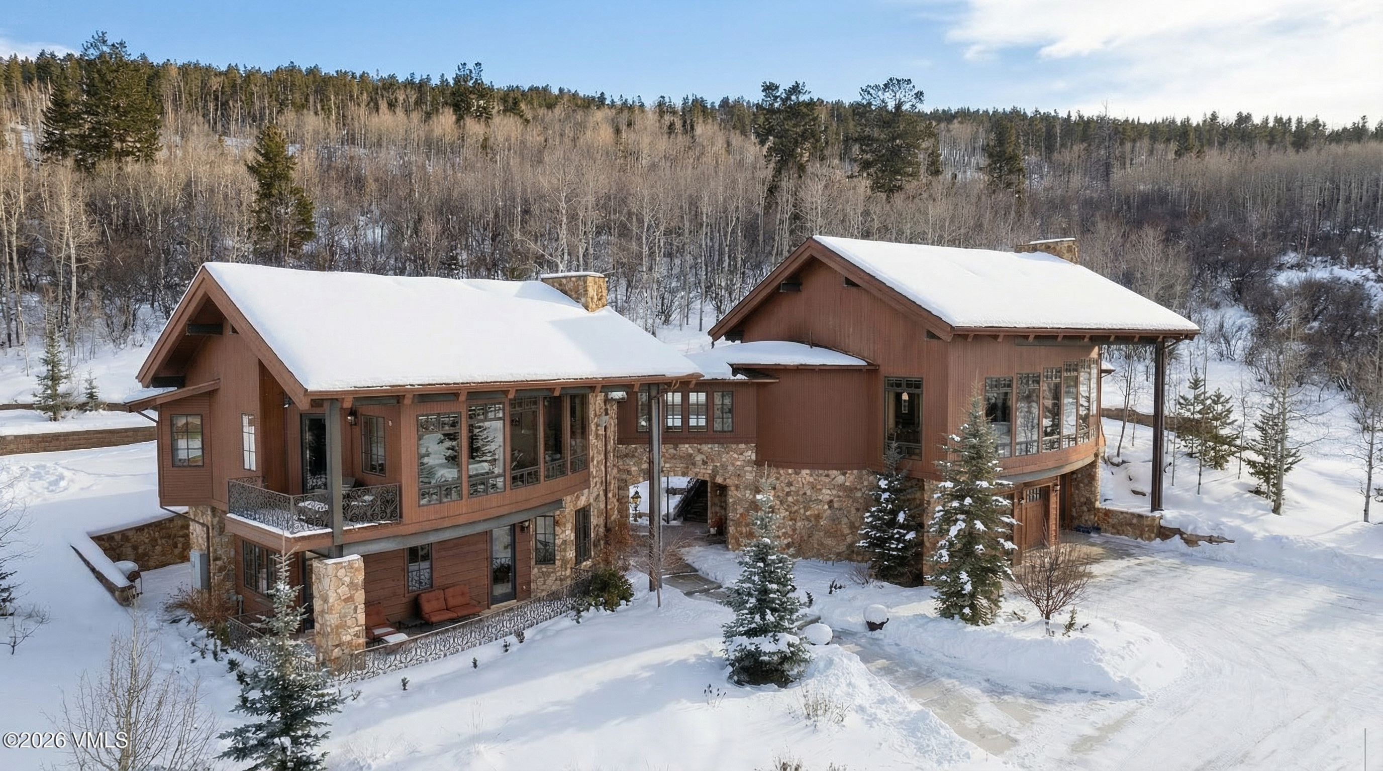 This is an exterior front view of a luxurious mountain home covered in snow. The architecture features a blend of wood and stone, with large windows offering stunning views. A stone archway connects two sections of the home, and the professionally landscaped yard adds to the property's charm.