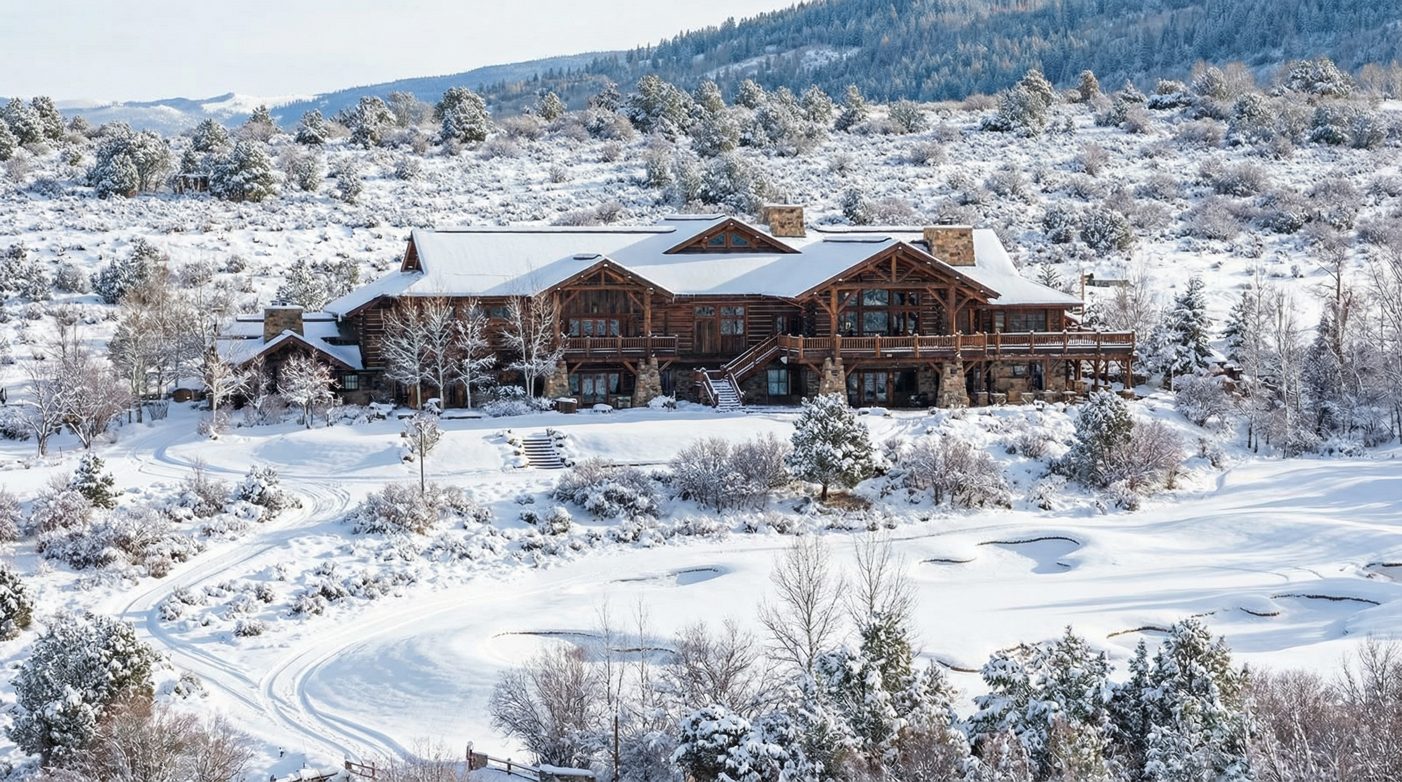This image showcases the front view of a grand log home nestled in a snowy, mountainous landscape. The architectural style exudes rustic luxury with its exposed timber beams, stone accents, and multiple windows. The property is surrounded by snow-covered trees and terrain, emphasizing its secluded and picturesque setting.